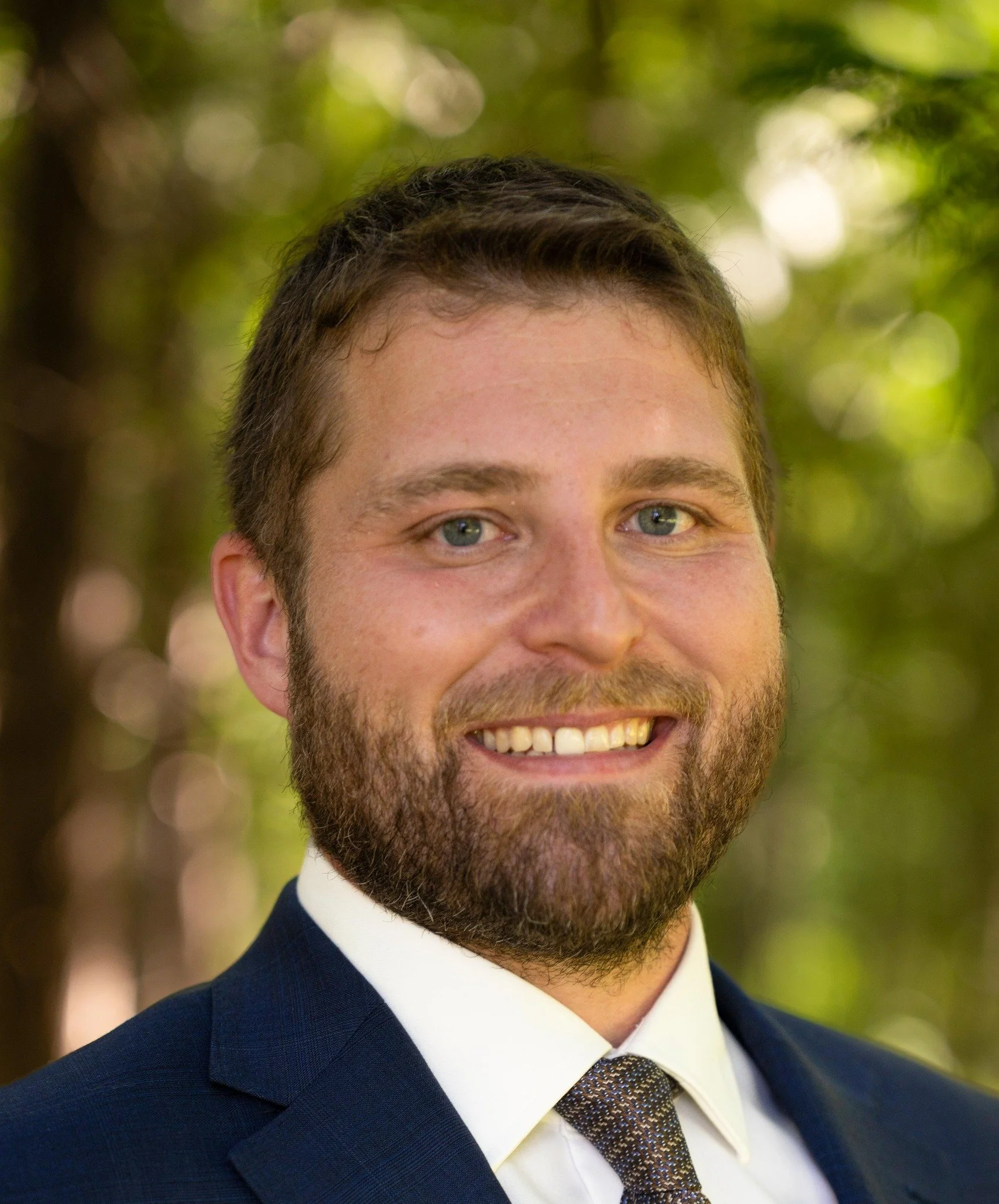 A smiling man with a beard and short hair, wearing a suit and tie, posing outdoors with a blurred green background.