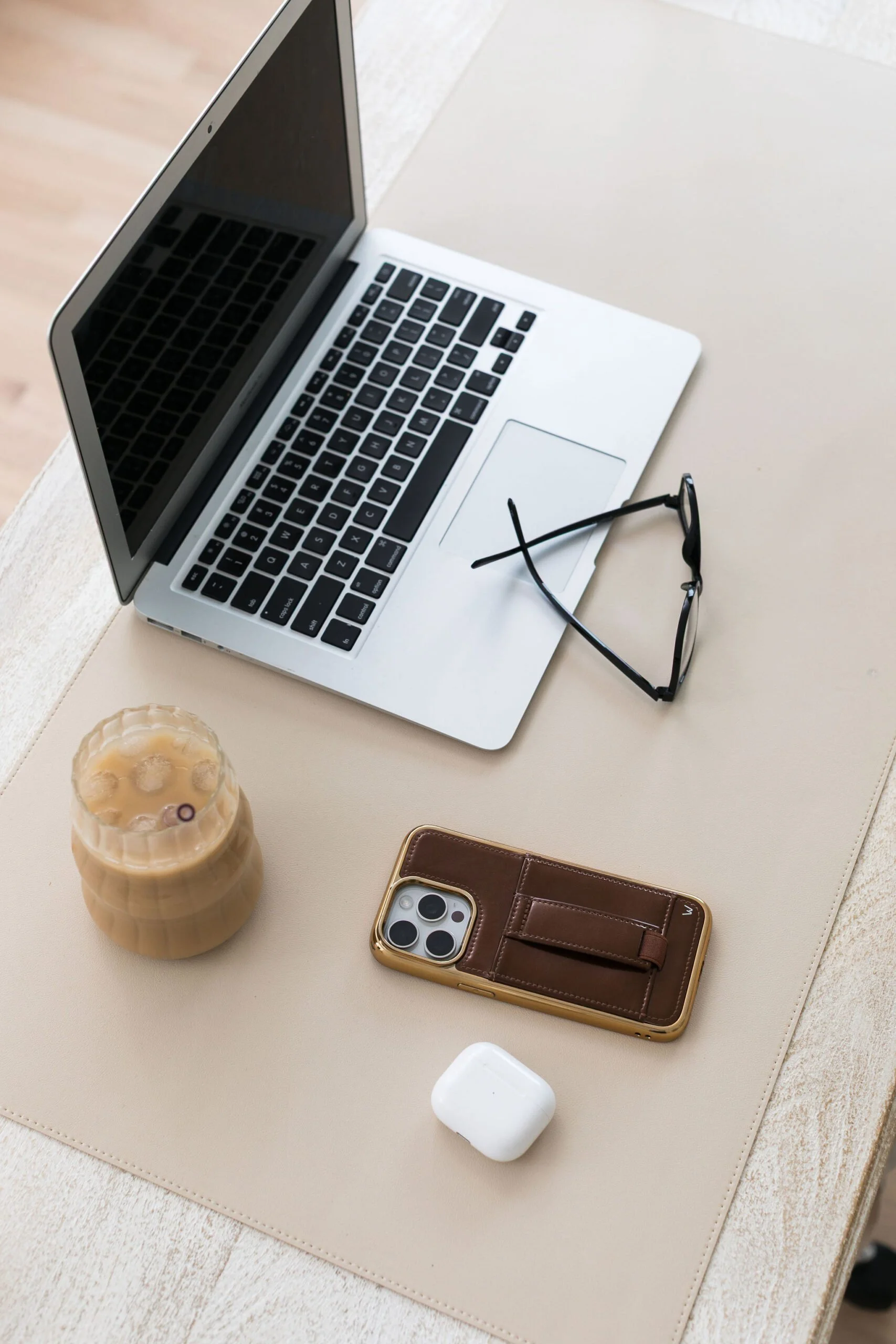 A laptop, glasses, a coffee drink, AirPods, a smartphone in a wallet case, and a small white object on a beige surface.