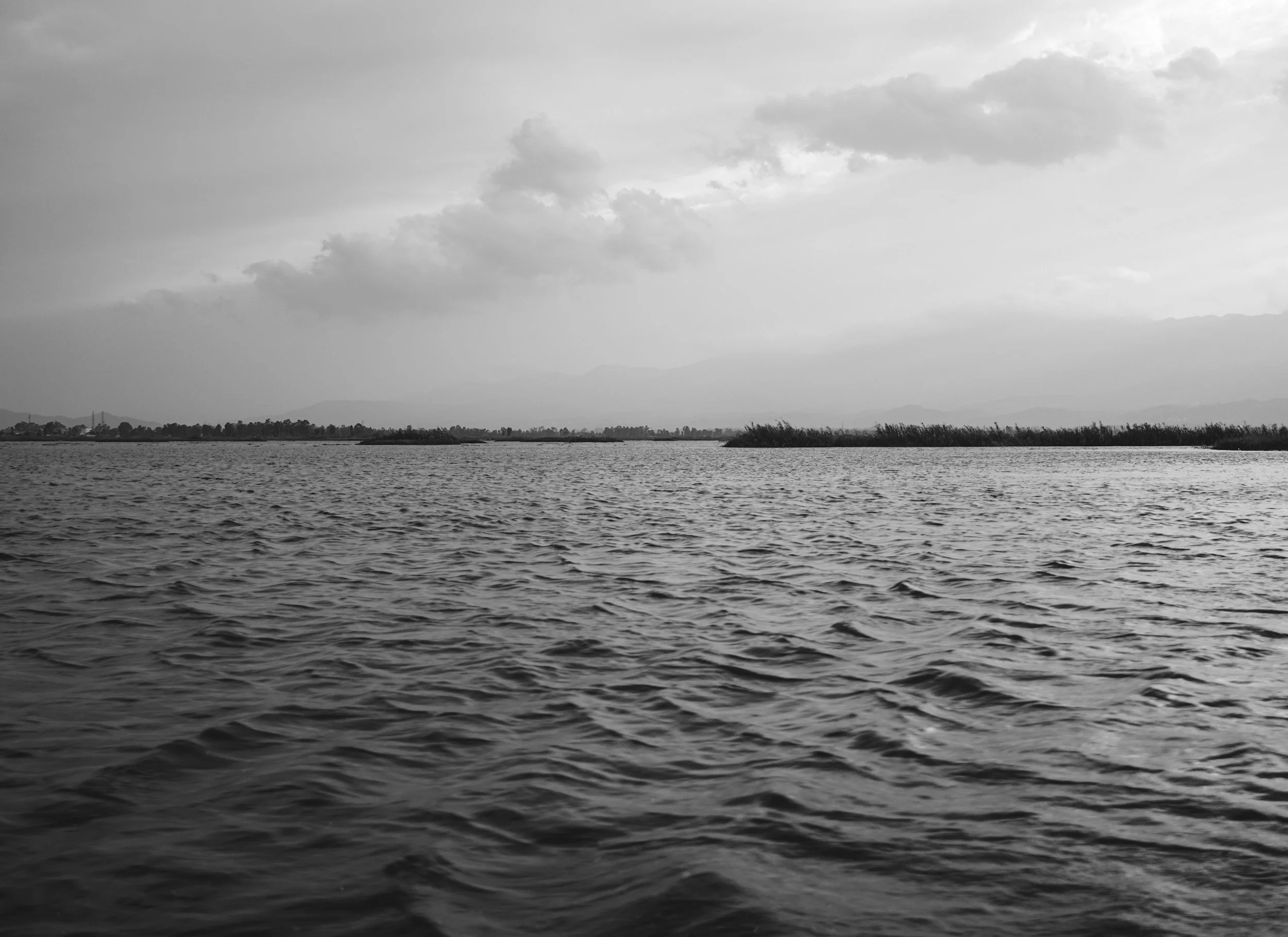 A black-and-white photo of a river with small ripples, with trees along the far bank and mountains in the distance under cloudy skies.