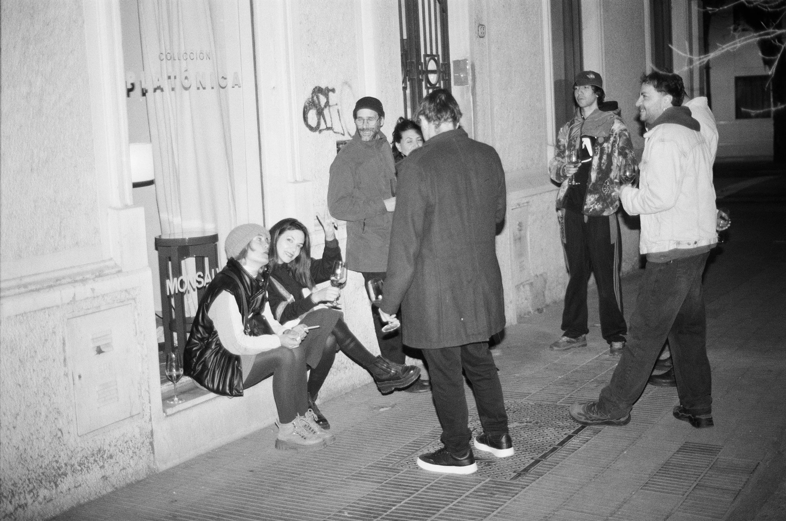Grupo de personas conversando en una reunión nocturna sobre una vereda, en una fotografía en blanco y negro en la Exposicion de Colección Platónica en 2025 en el estudio showroom
