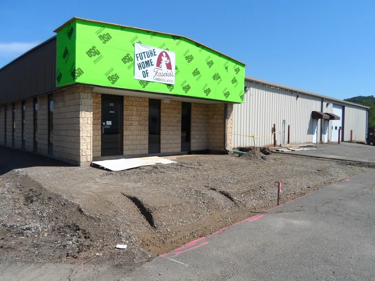 The exterior of a building under construction with a green sign that says "Future Home of Faccia's Chocolates." The building has brick and metal siding with some construction materials and dirt in front.