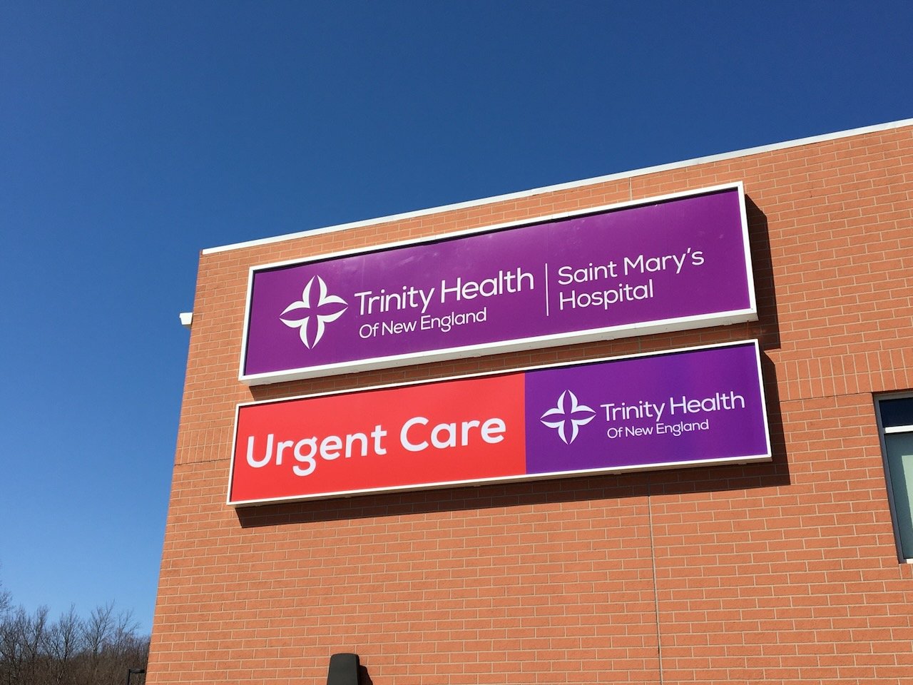 Signs on the exterior of a brick hospital building for Trinity Health of New England and Saint Mary's Hospital, with an urgent care sign in red, indicating the hospital provides urgent care services.