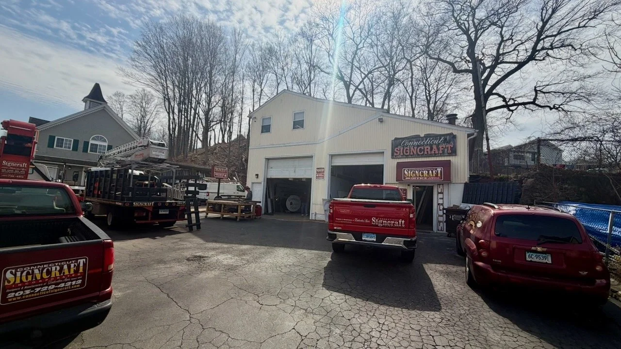 A building with a sign that reads 'Connecticut Signcraft' on the front, with several trucks and cars parked outside, and a few leafless trees in the background.