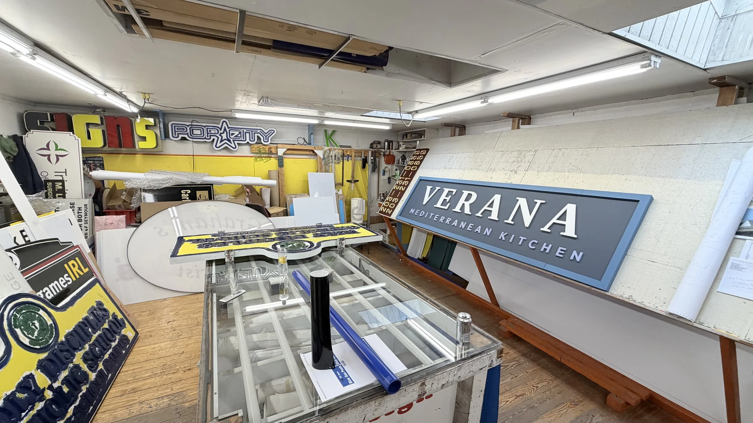 Workshop with various signage and sign frames, including a prominent large sign for 'VERANA MEDITERRANEAN KITCHEN' leaning against the wall, tools and materials scattered around, and bright fluorescent lighting.