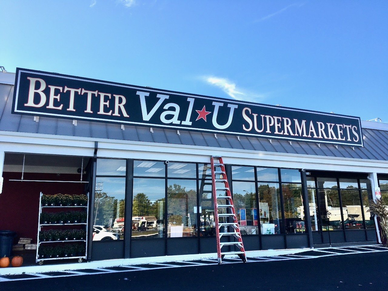 Supermarket storefront with a large sign that reads 'Better Val*U Supermarkets' above the entrance, with a ladder leaning against the building and pumpkins outside.