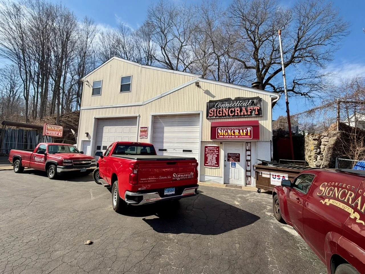A building with signs for Connecticut Signcraft, with three red vehicles parked in front, including a pickup truck and a van, on a cracked asphalt lot. The building has three garage doors and is surrounded by trees without leaves.