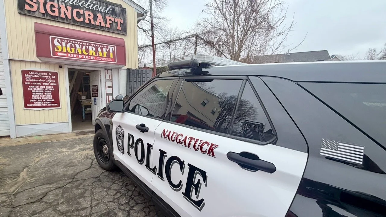 A police vehicle parked outside a sign craft shop with signs indicating custom signs and designs in the background.