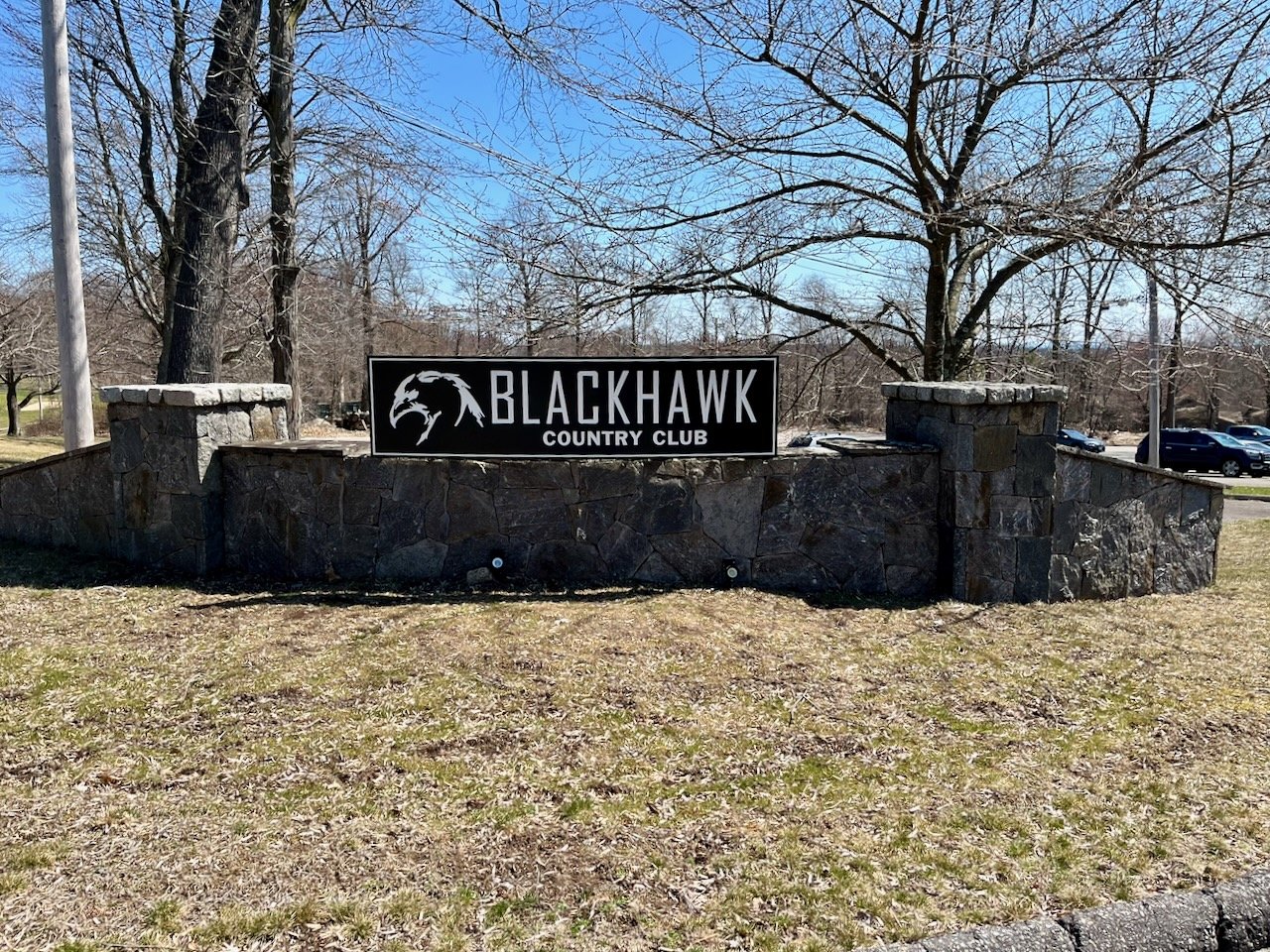 Sign for Blackhawk Country Club on a stone wall with trees and parked cars in the background.