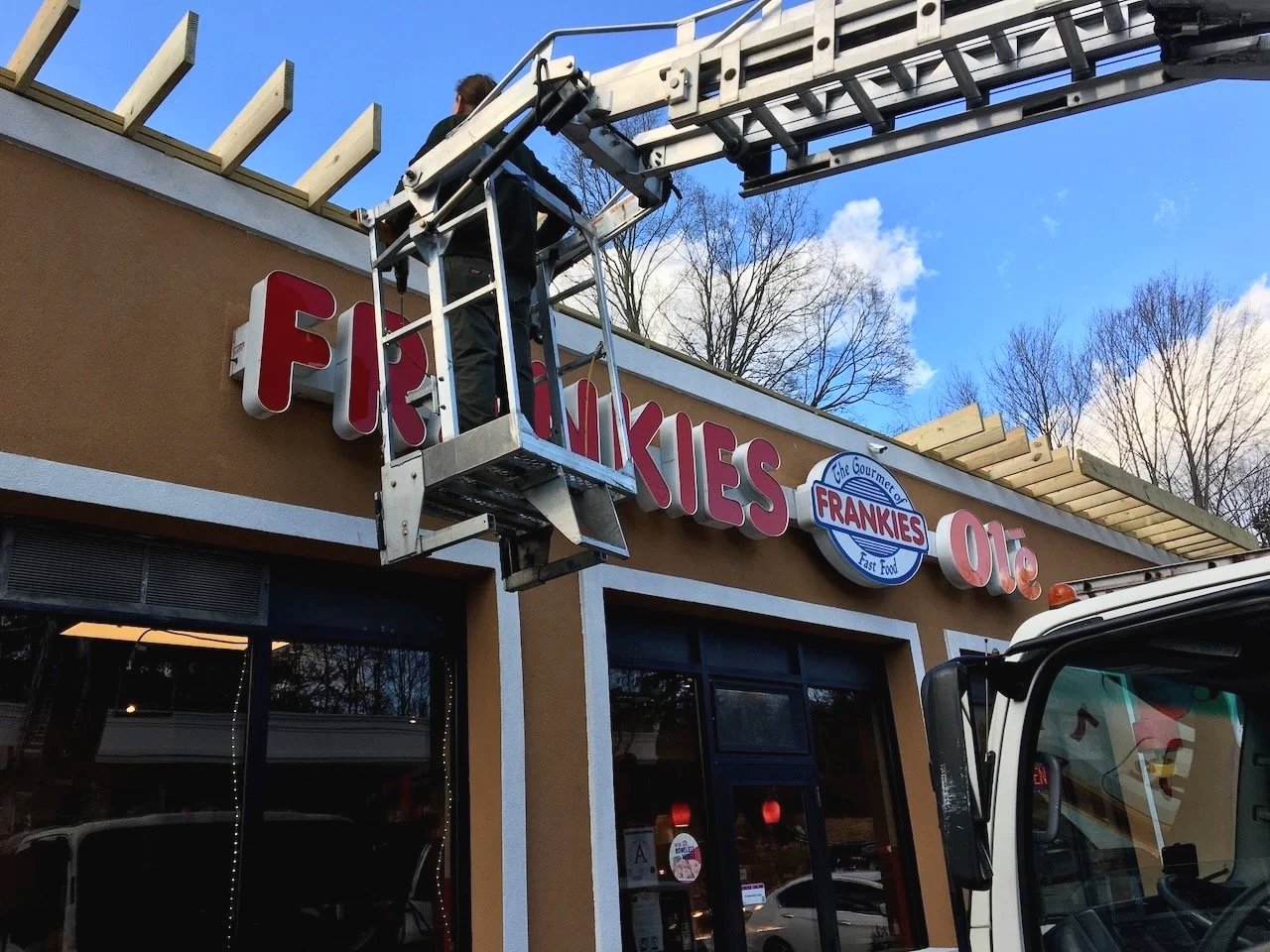 A worker on a lift installing or repairing a sign for Frankie's restaurant on the building facade. The sign reading 'FRANKIES' is partially visible, with a decorative logo in the center.