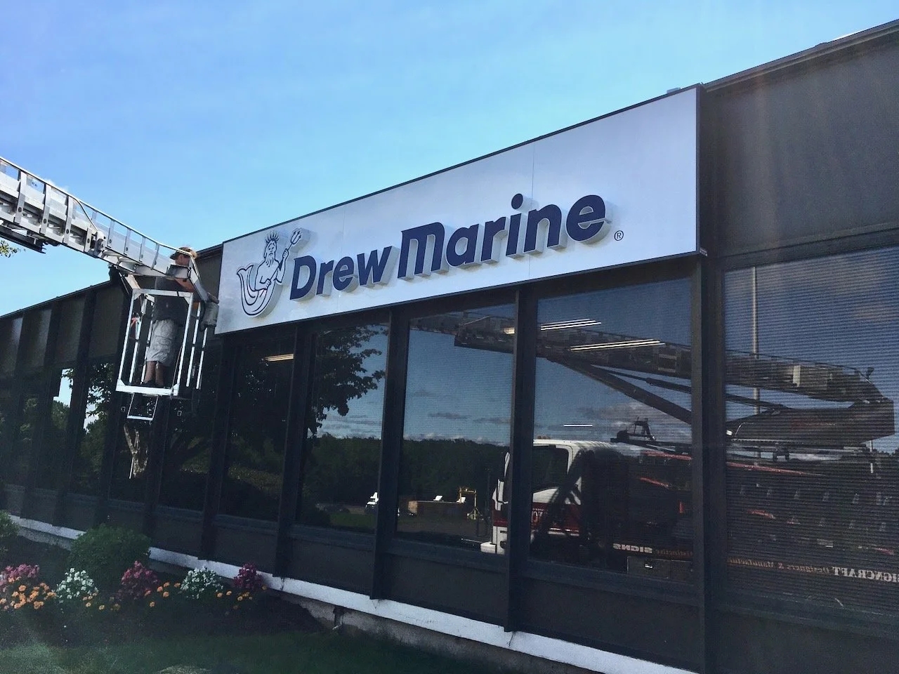 A person installing or maintaining the Drew Marine store sign using a lift, with the store's glass front and flower bed visible.