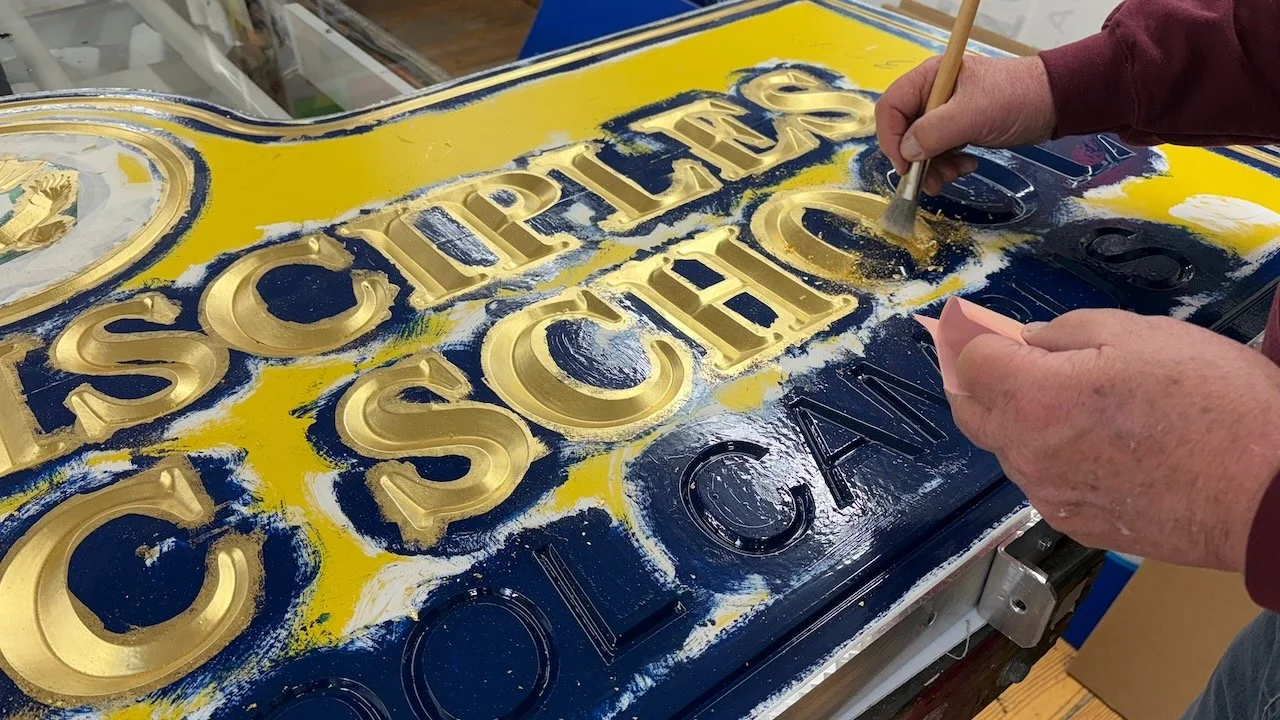 A person using a brush to apply gold paint or gilding onto a vintage sign with raised letters, on a work table.