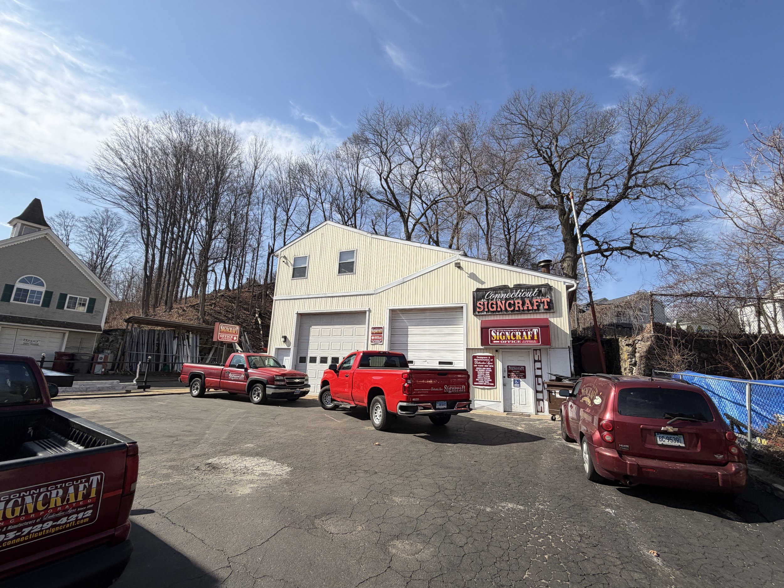 A parking lot in front of a beige building with signs that read 'Connecticut Signcraft' and several red trucks parked nearby. Behind the building, there are leafless trees and a clear blue sky.