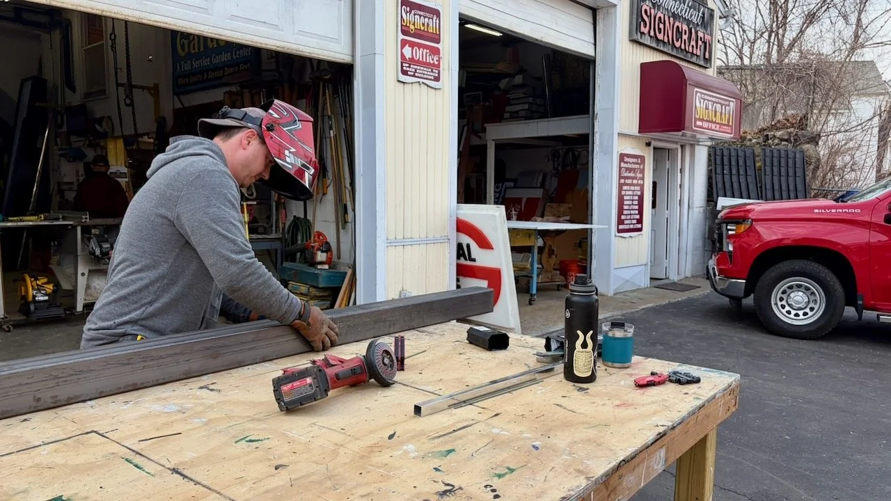 A person wearing a gray hoodie, gloves, and a red helmet is working with a piece of wood or metal at a woodworking or metalworking shop outside a building labeled Connecticut Signcraft. The person is focused on the work, and tools like a cordless drill, a square, and safety glasses are on the worktable.