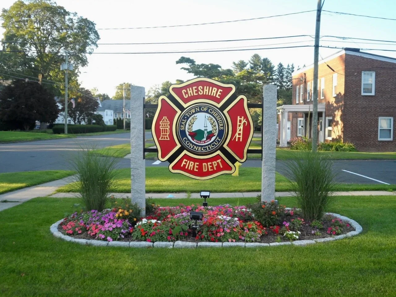Sign for Cheshire Fire Department in Connecticut, featuring a fire department emblem with a lighthouse and bridge, surrounded by flowers in a landscaped area.