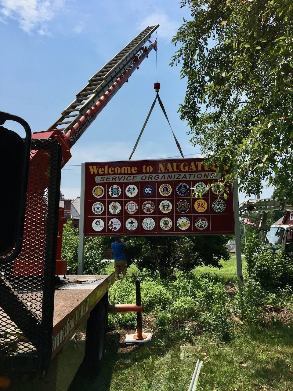 A fire truck with an extended ladder lifting a large sign that says "Welcome to NAUGAT" and features various service organization logos. A person in a blue shirt and khakis stands below, and there are trees and greenery around.