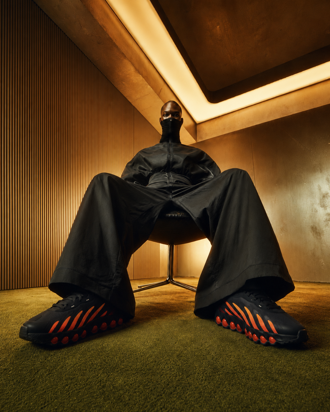 A person sitting on a chair in a modern room with warm lighting, wearing black athletic shoes with orange accents, wide-leg black pants, a black jacket with a high collar, and a black face covering, viewed from a low angle.