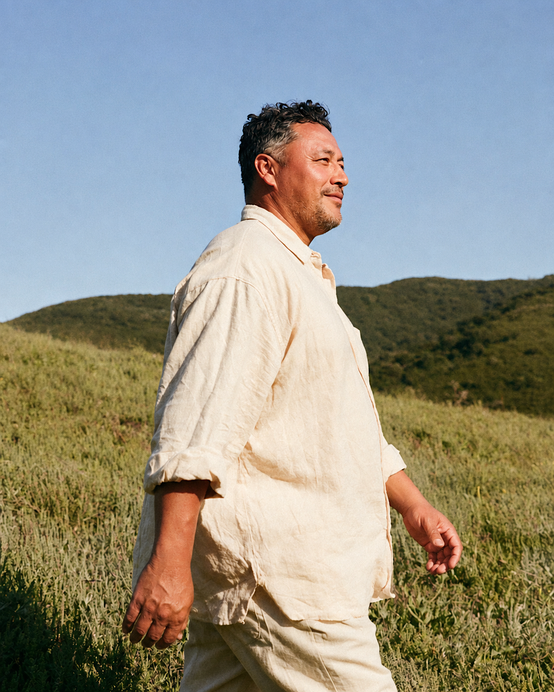 A middle-aged man walking outdoors in a grassy field with rolling hills in the background, wearing a light-colored long-sleeve shirt and pants, with the sun shining on his face. IDK Agency