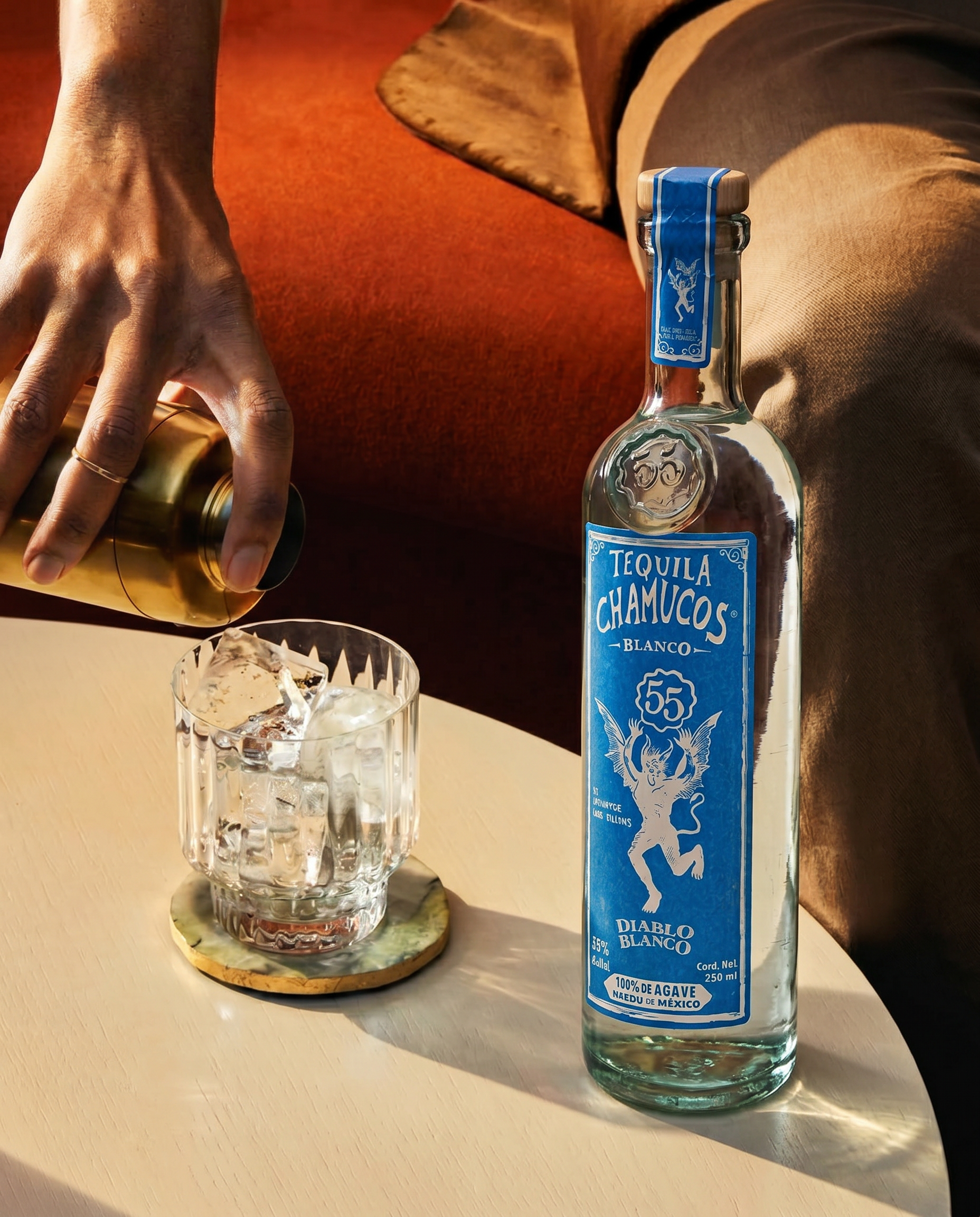 A glass of ice and clear liquid on a coaster, a bottle of tequila chamucos blanco with a blue label, a hand reaching for a gold-colored can or bottle, and a person wearing beige and brown clothing sitting on a red chair in the background. IDK Agency