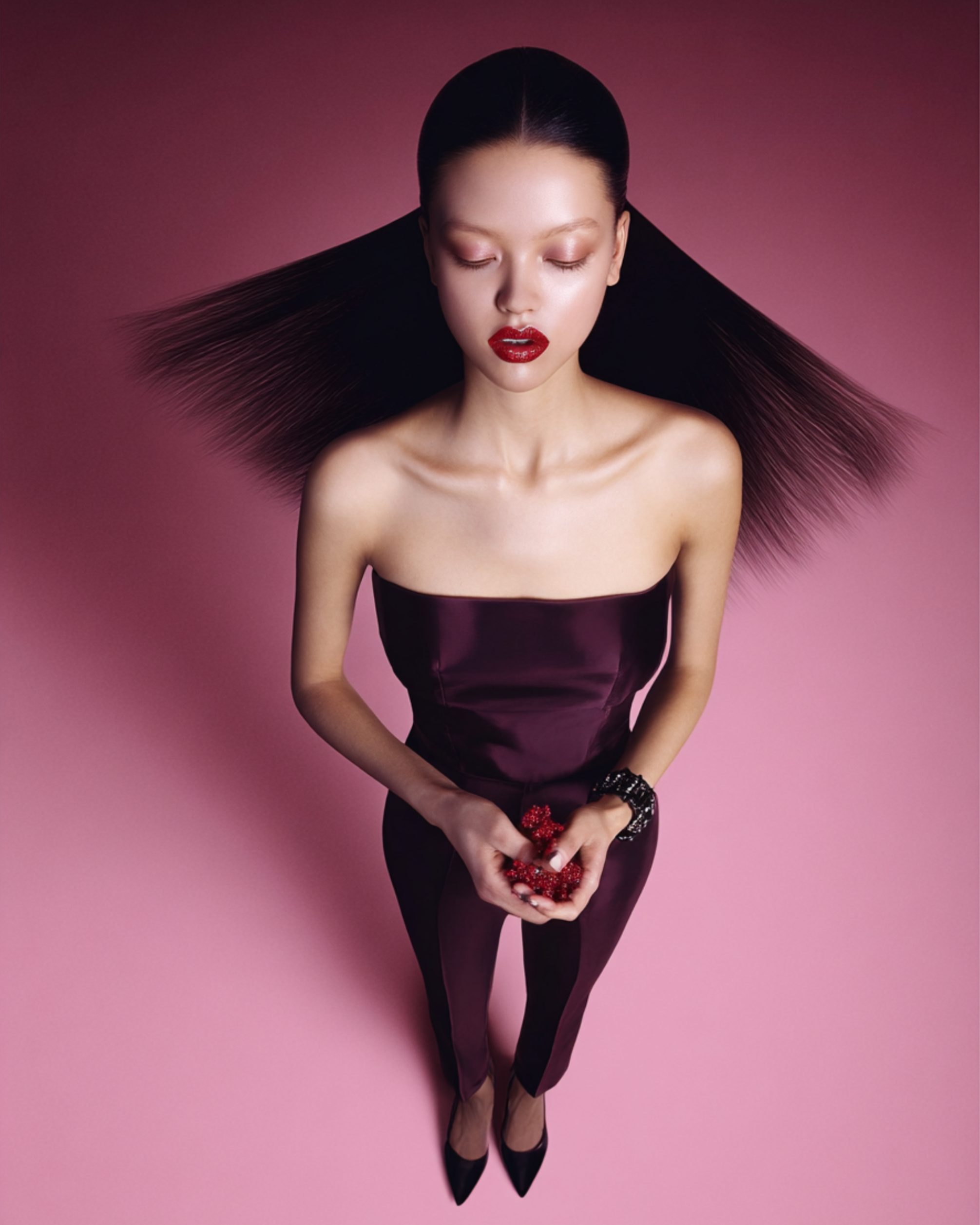 idk agency A woman with long, straight black hair, closed eyes, and red lipstick, standing against a pink background. She wears a strapless dark purple outfit, holding red beads in her hands, and has a bracelet on her wrist.