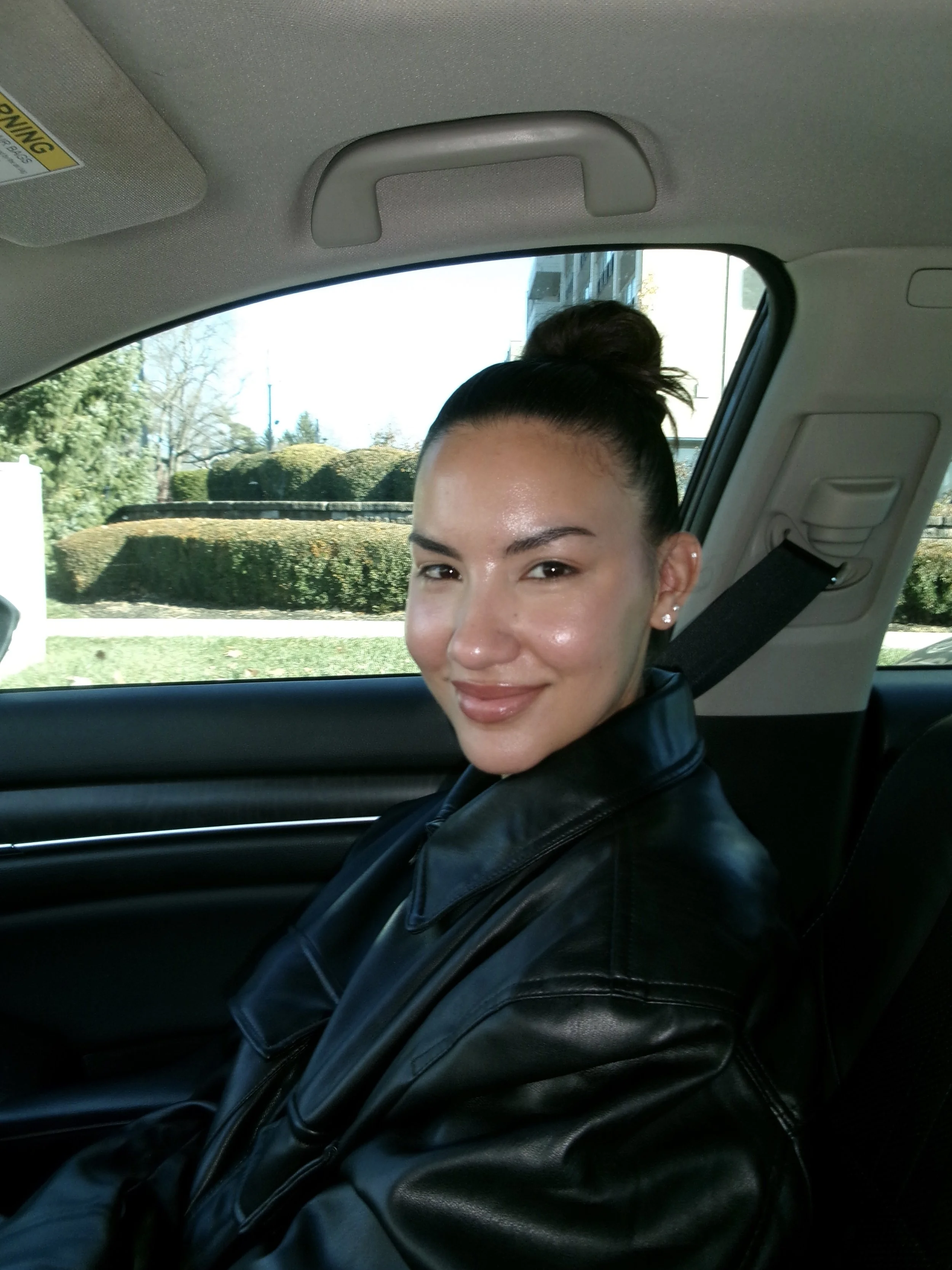 A woman with a high bun hairstyle and pierced ears sitting in a car, wearing a black leather jacket, smiling with a background of greenery outside a window.