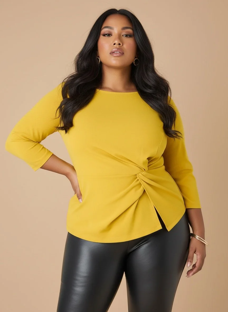 A confident woman with long, dark hair and makeup, wearing a bright yellow top with a twist knot detail and black leather pants, standing against a beige background.