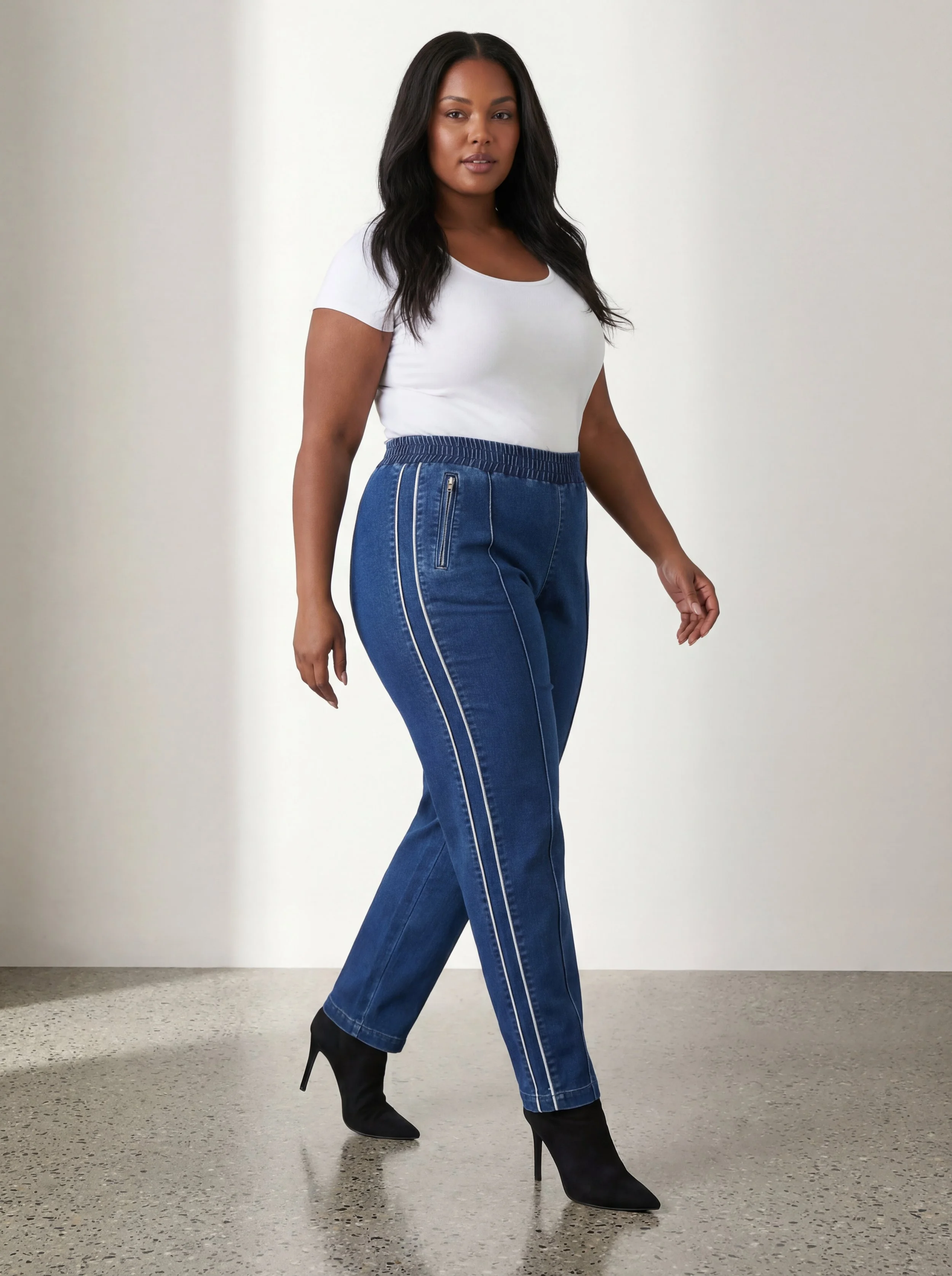 A woman walking in black high heel boots, wearing a white t-shirt and blue jeans with white side stripes.