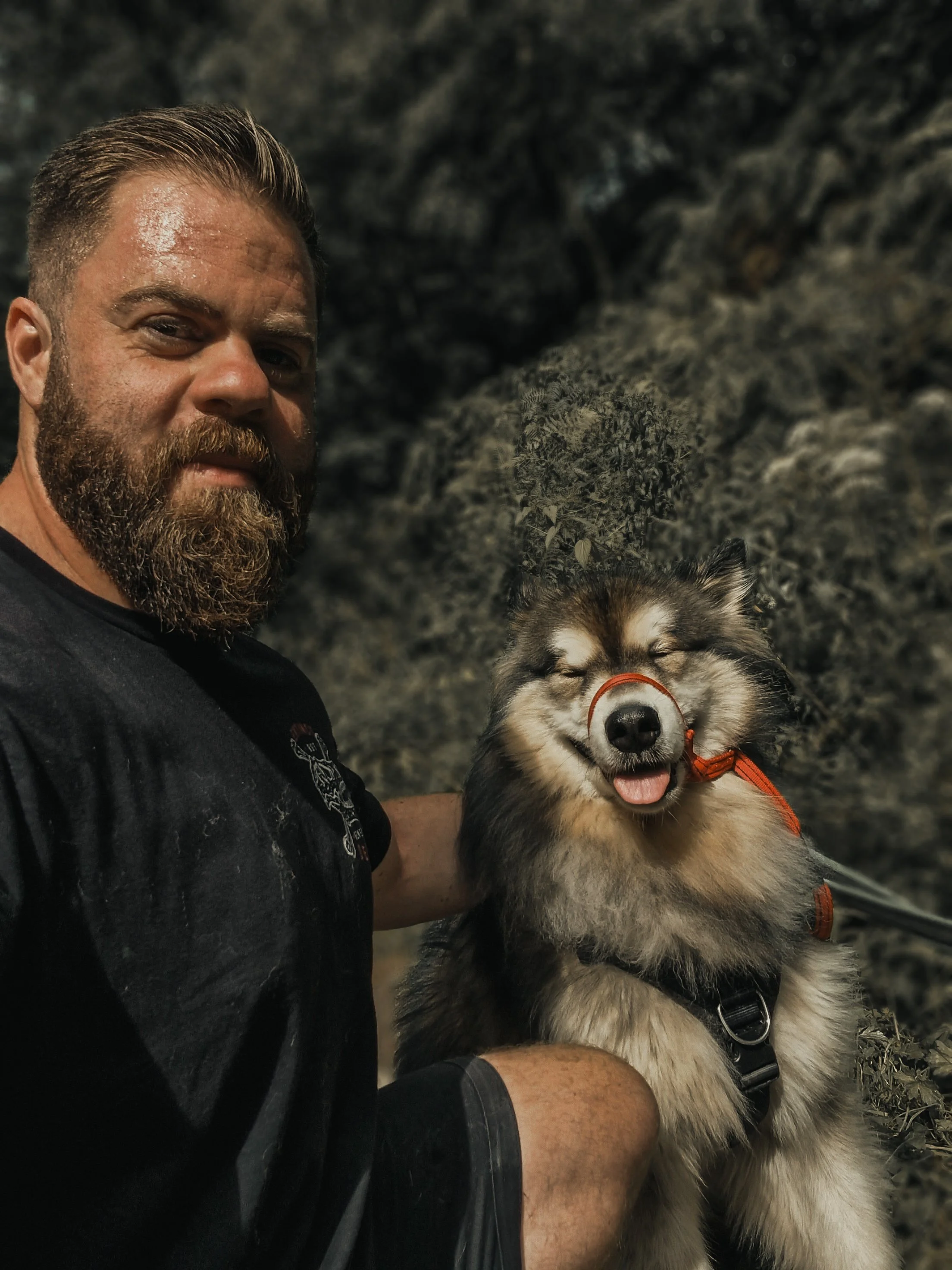 A man with a beard and mustache taking a selfie with a fluffy Husky dog outdoors against a rocky background. The dog is smiling with eyes closed and tongue slightly out.