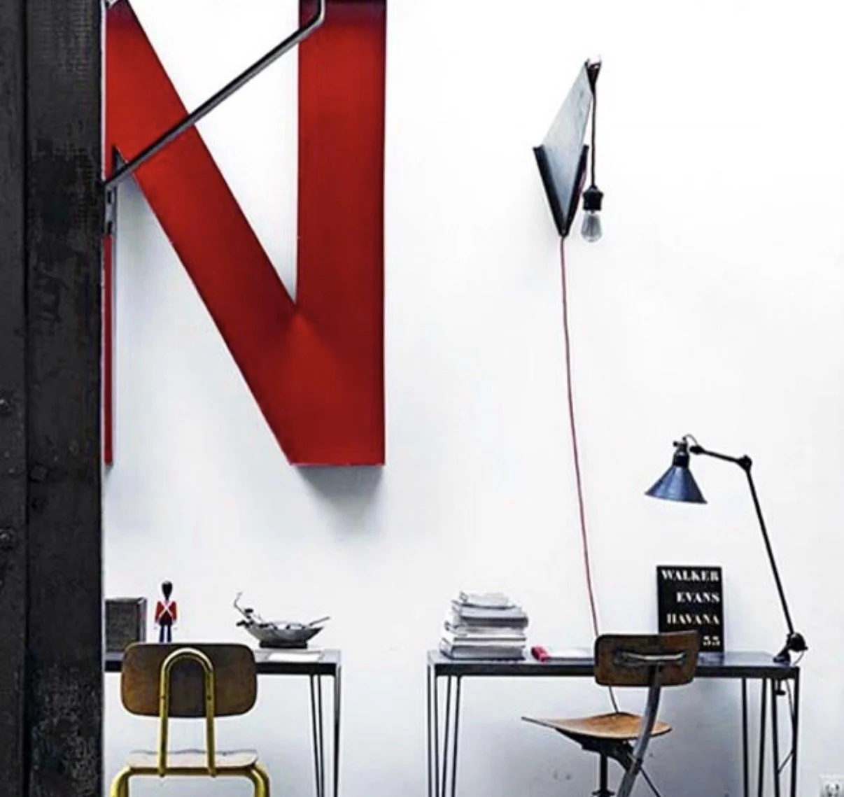 Interior workspace with two desks, one with a wooden chair with yellow metal legs and the other with a wooden chair. The left desk has a small figure and a bowl, the right desk has stacks of papers and a sign that reads 'Walker Evans Blurbs.' A large red letter 'N' is mounted on the wall, with a black and silver wall lamp, a small wall-mounted lamp, and a hanging light bulb with a red cord.