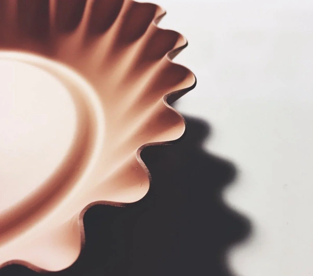 A close-up of a copper-colored, scalloped-edged baking tin, casting a shadow on a white surface.