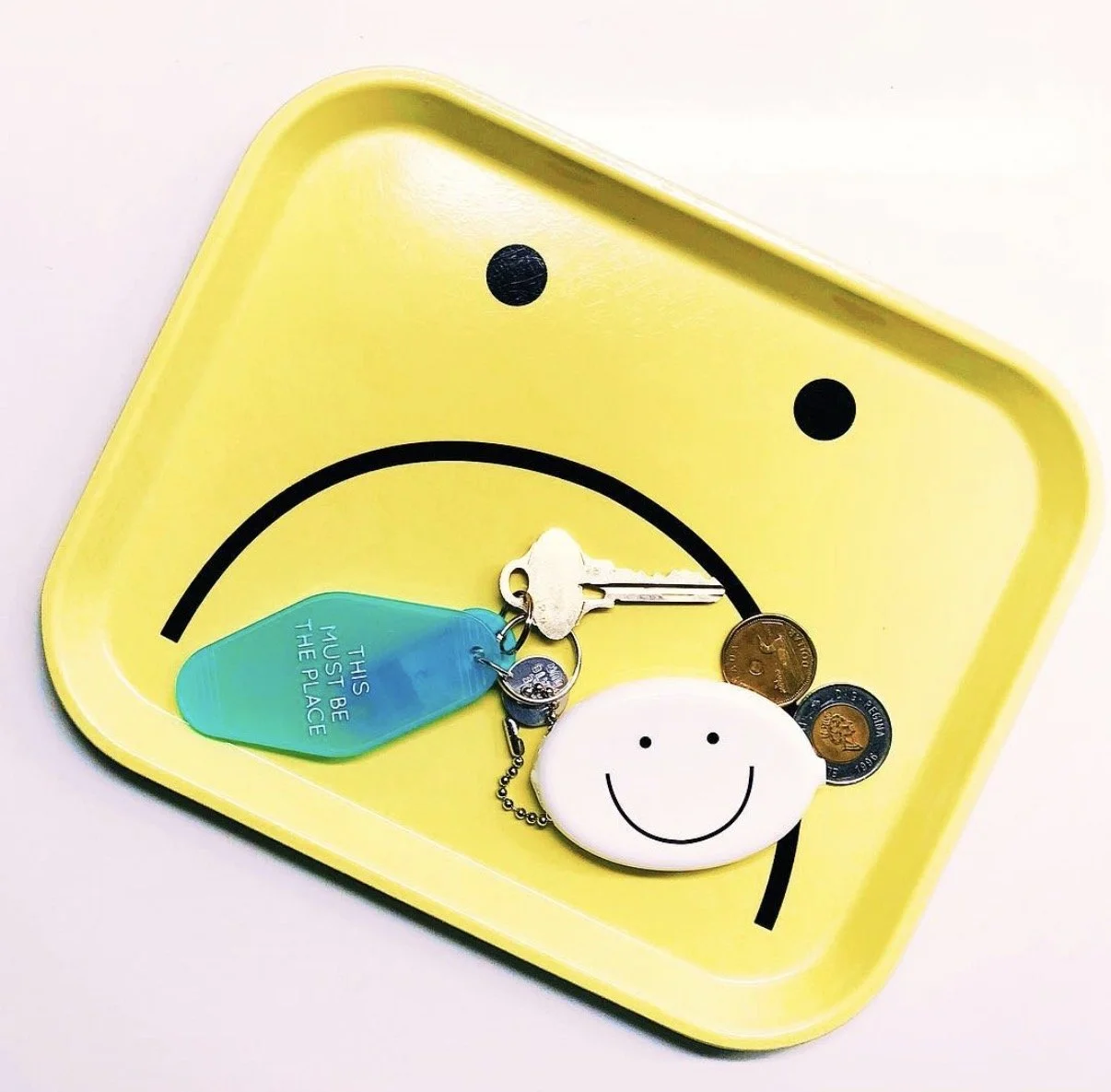 A yellow tray with a sad face printed on it, holding keys, coins, and a smiley face keychain and tag.