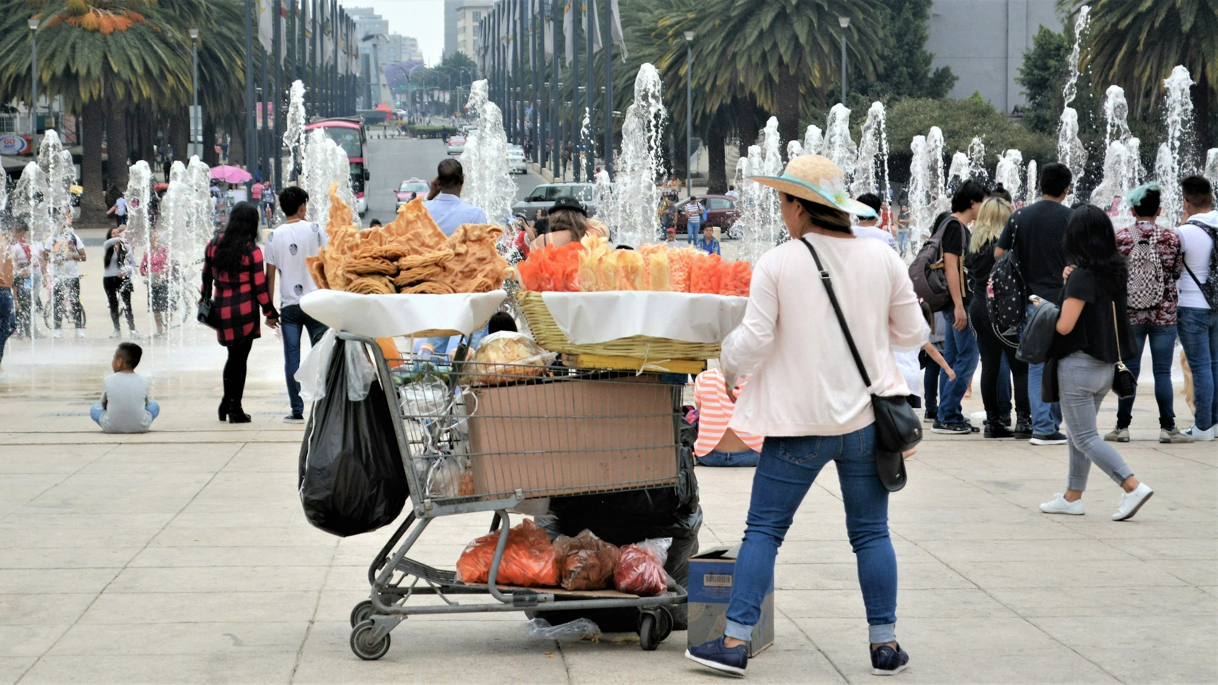 A busy urban scene with a woman in a white shirt and blue jeans browsing street food in front of a cart, surrounded by people near two water fountains, trees, cars, and buildings in the background.