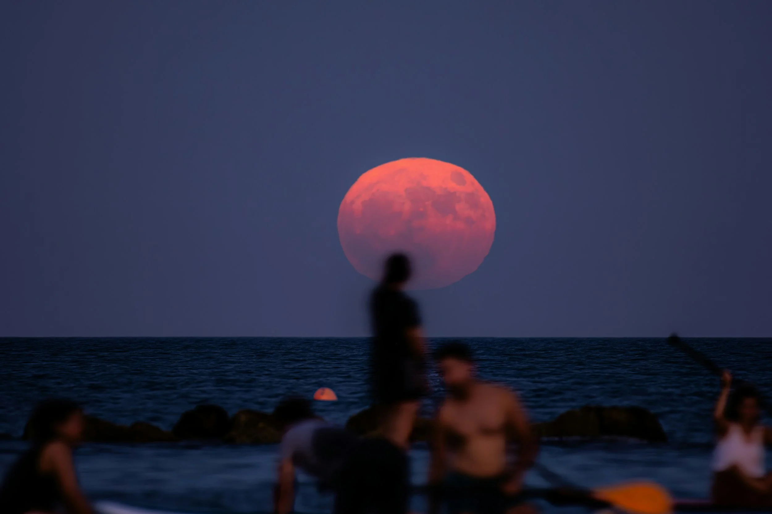 People gathered on a beach at sunset with the moon rising over the ocean, creating a large reddish moon in the sky.