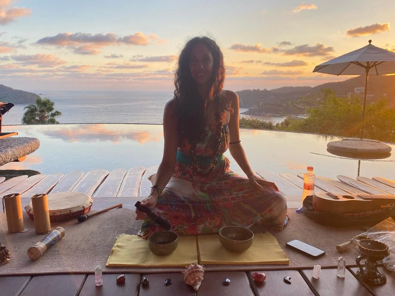 A woman sitting cross-legged on a mat, performing meditation or a spiritual ritual outdoors during sunset, with a view of the ocean and hills in the background. She is surrounded by ritual objects, bowls, crystals, and a water bottle.