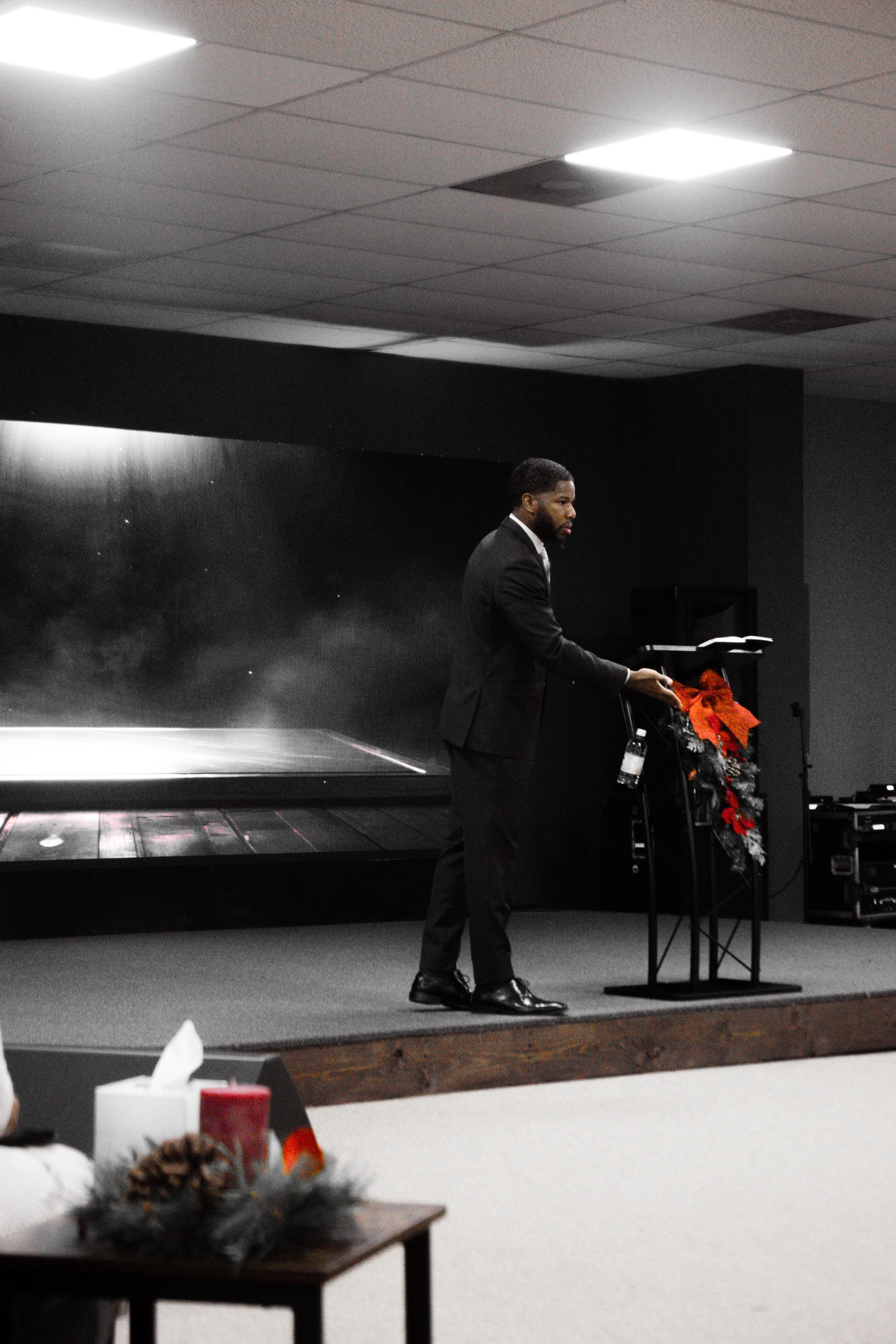 A man in a black suit stands at a podium decorated with Christmas ornaments, speaking or delivering a speech in a room with a dark background and a large screen behind him.
