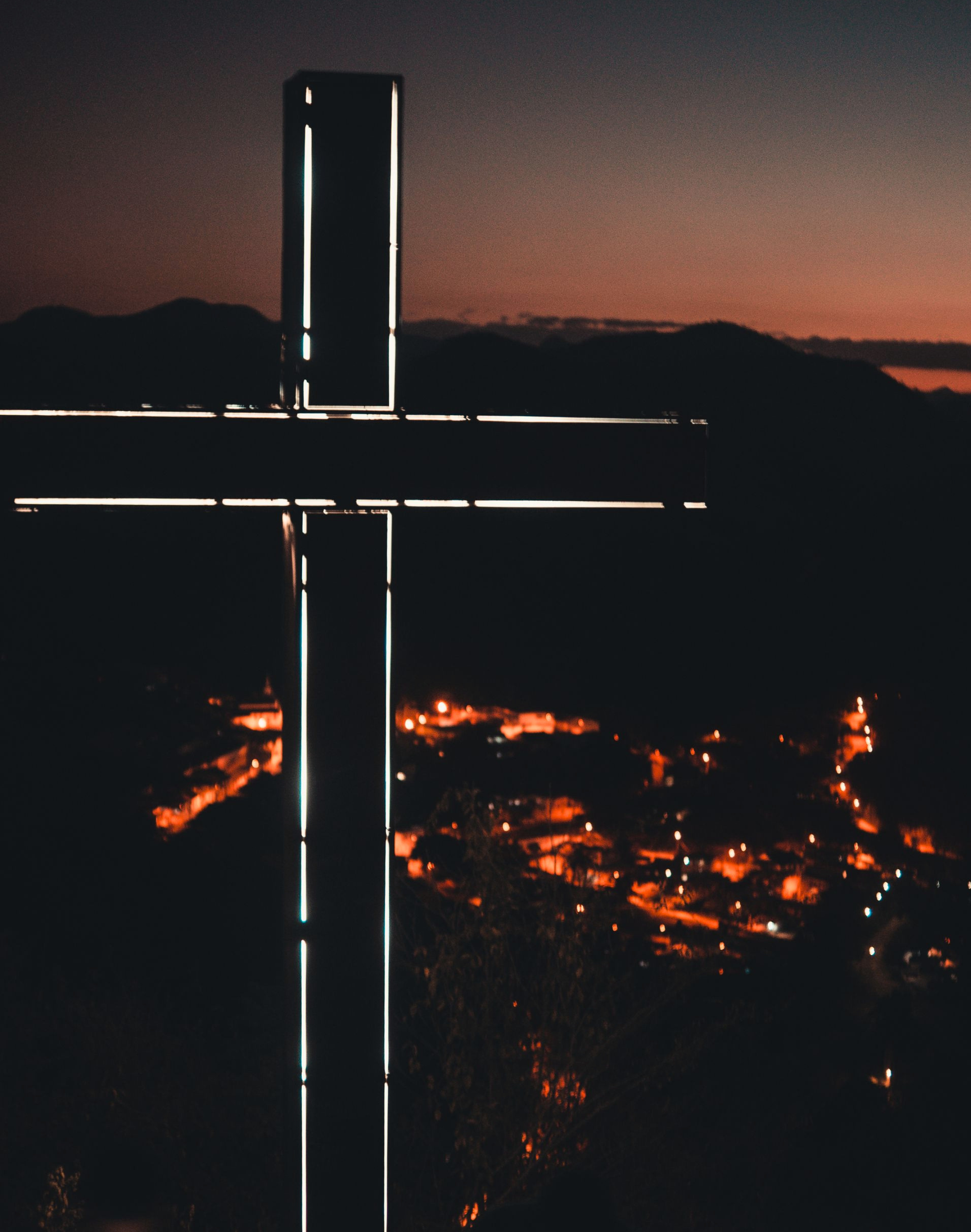 A landscape scene at dusk or dawn with a silhouette of mountains in the background, a dark sky with orange and pink hues near the horizon, illuminated city lights below, and a structure with vertical and horizontal neon-like white lights in the foreground.