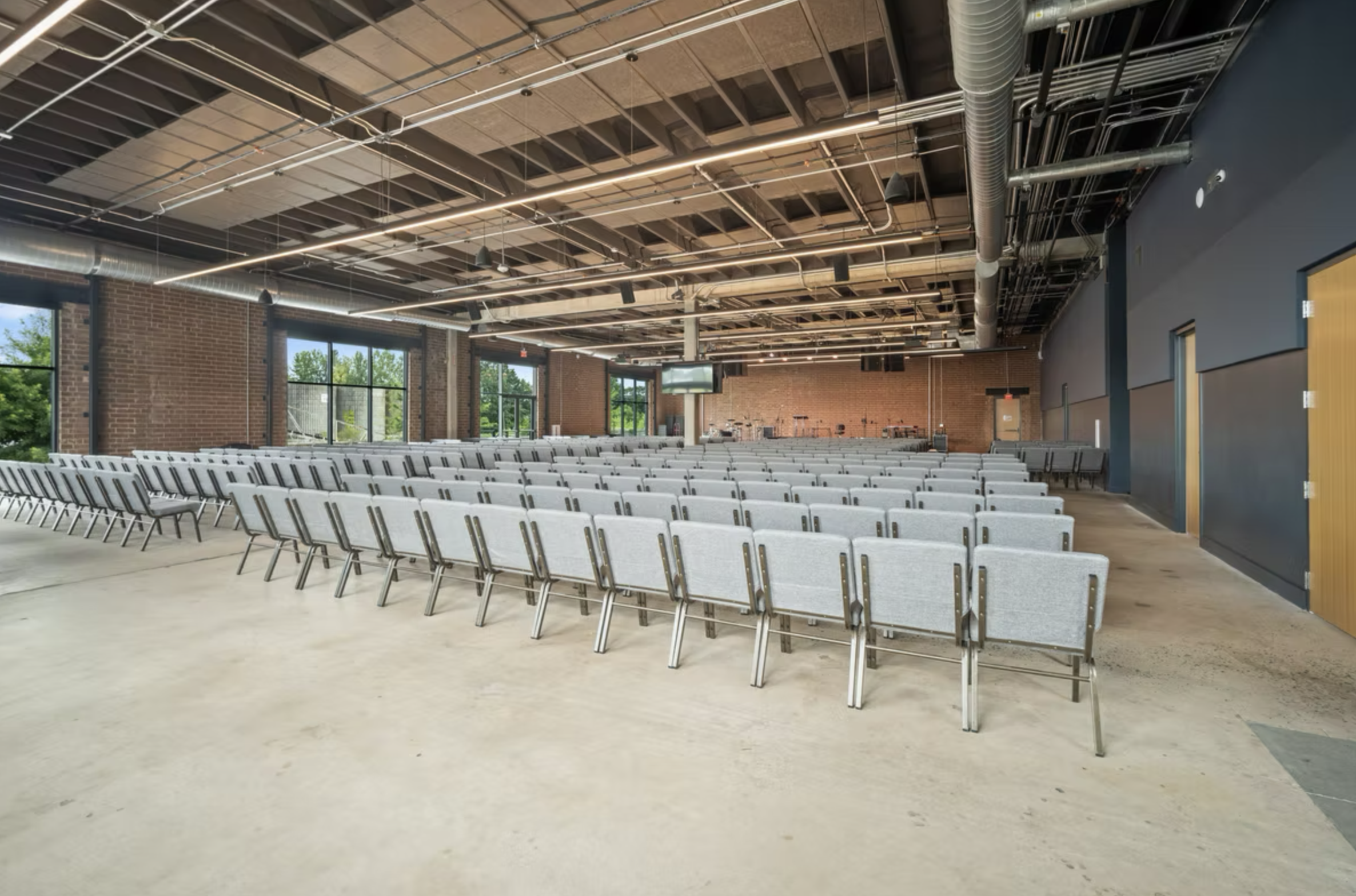 An empty modern auditorium with rows of gray chairs, large windows, exposed brick walls, and a high ceiling with visible industrial piping, lights, and a large screen at the front.