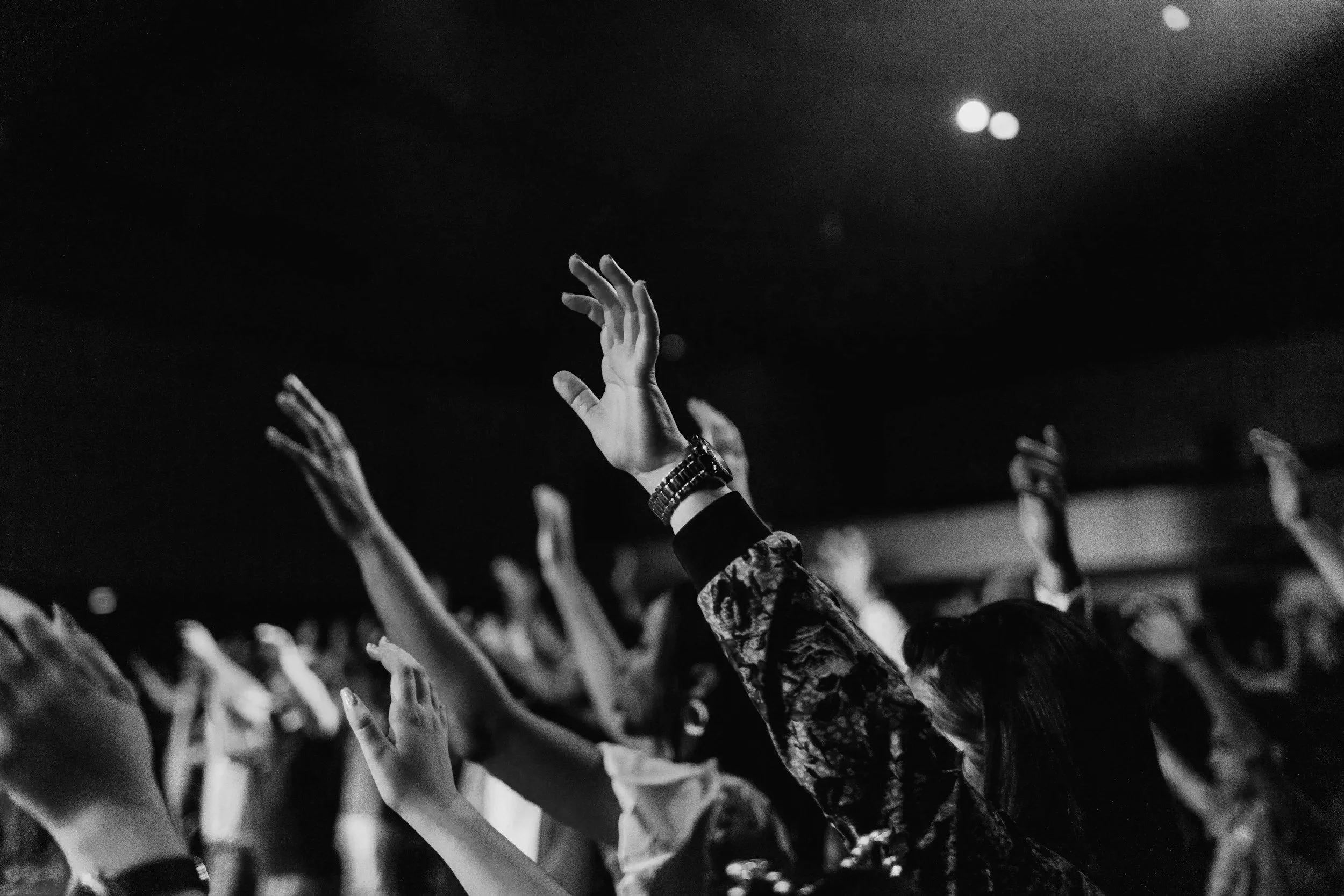 Hands raised during a concert or event, with some wearing watches or bracelets, in black and white.