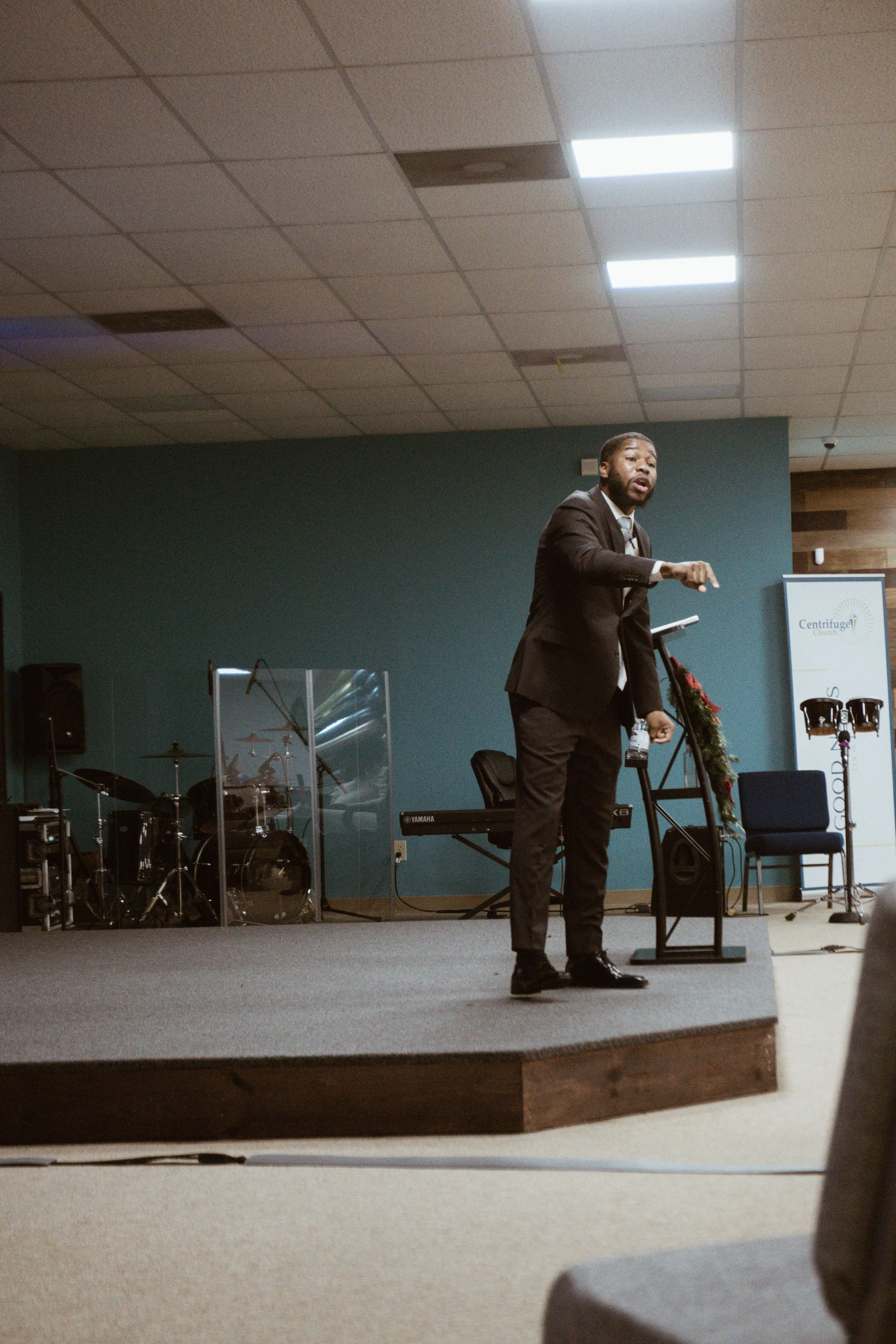 A man in a suit speaking on stage in a church or conference setting, with musical instruments and a sign in the background.