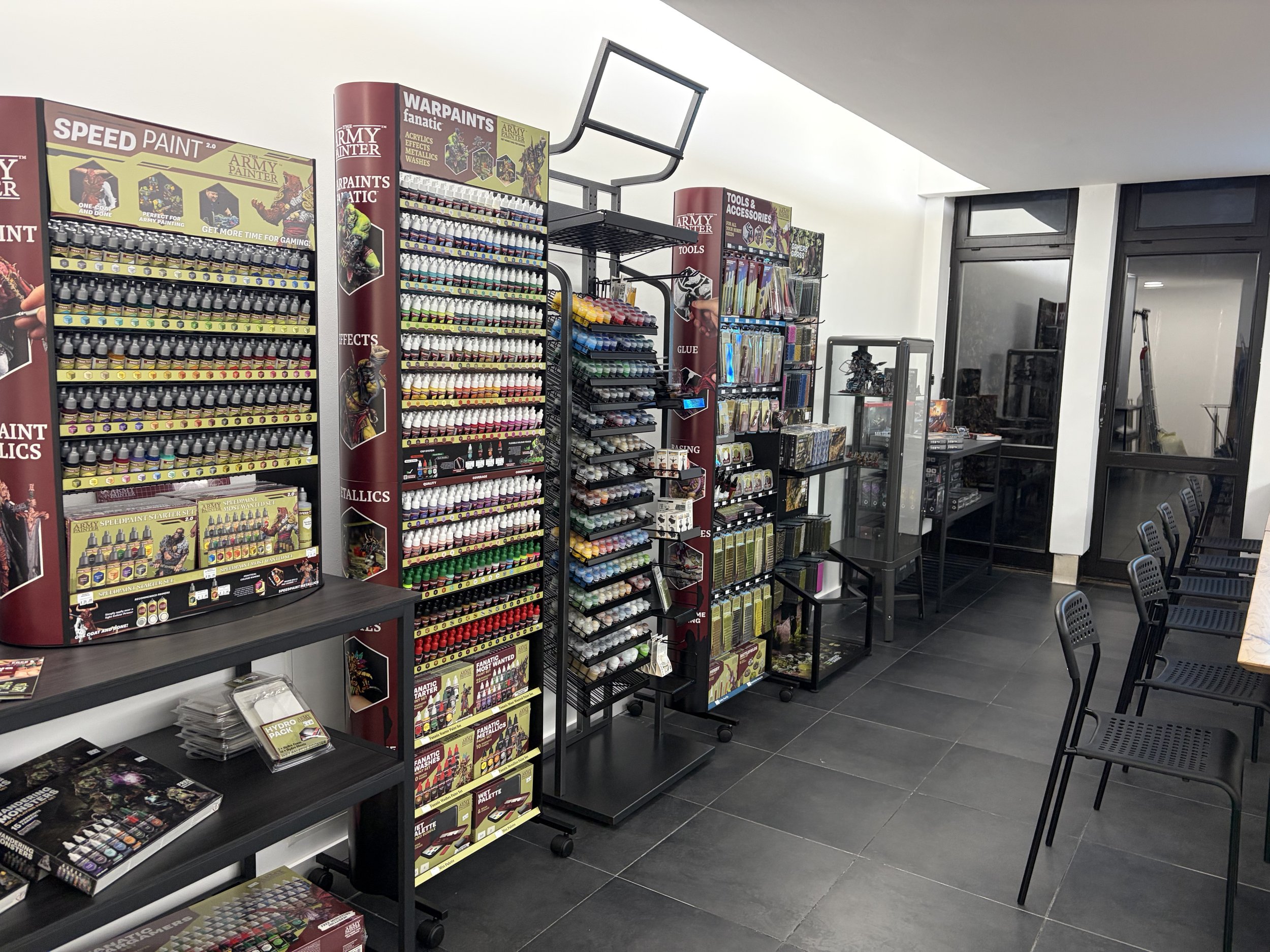 Regale mit Modellier- und Bastelmaterialien, darunter farbige Farben, Pinsel, Werkzeug- und Zubehörsets, in einem Geschäft. Im Hintergrund sind Tische und Stühle in einem Raum mit großen Fenstern.