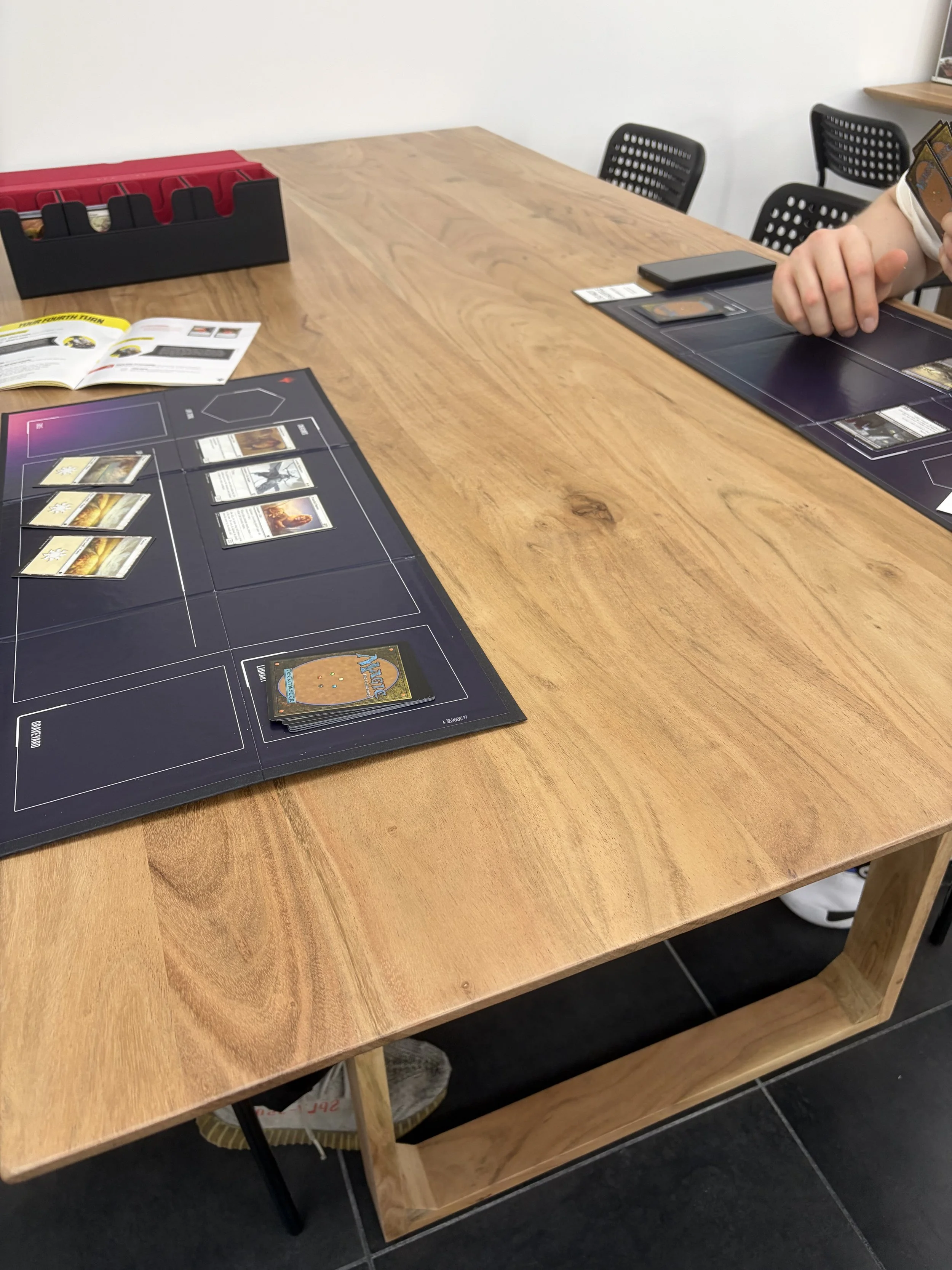 Ein Holztisch mit Karten und Spielmaterial für ein Kartenspiel, umgeben von schwarzen Stühlen.