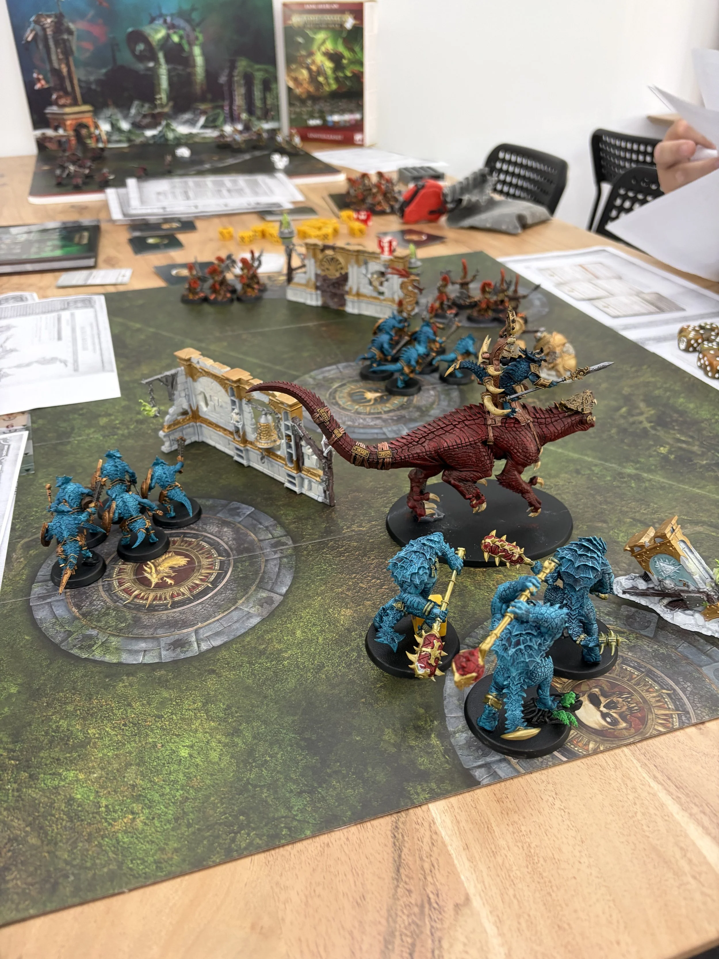Eine Tischplatte mit Miniaturfiguren für ein Brettspiel, darunter Drachen, Figuren in blauer Farbe, und ein Spielfeld mit Geländemerkmalen, sowie Spielmaterialien und Anleitungen.