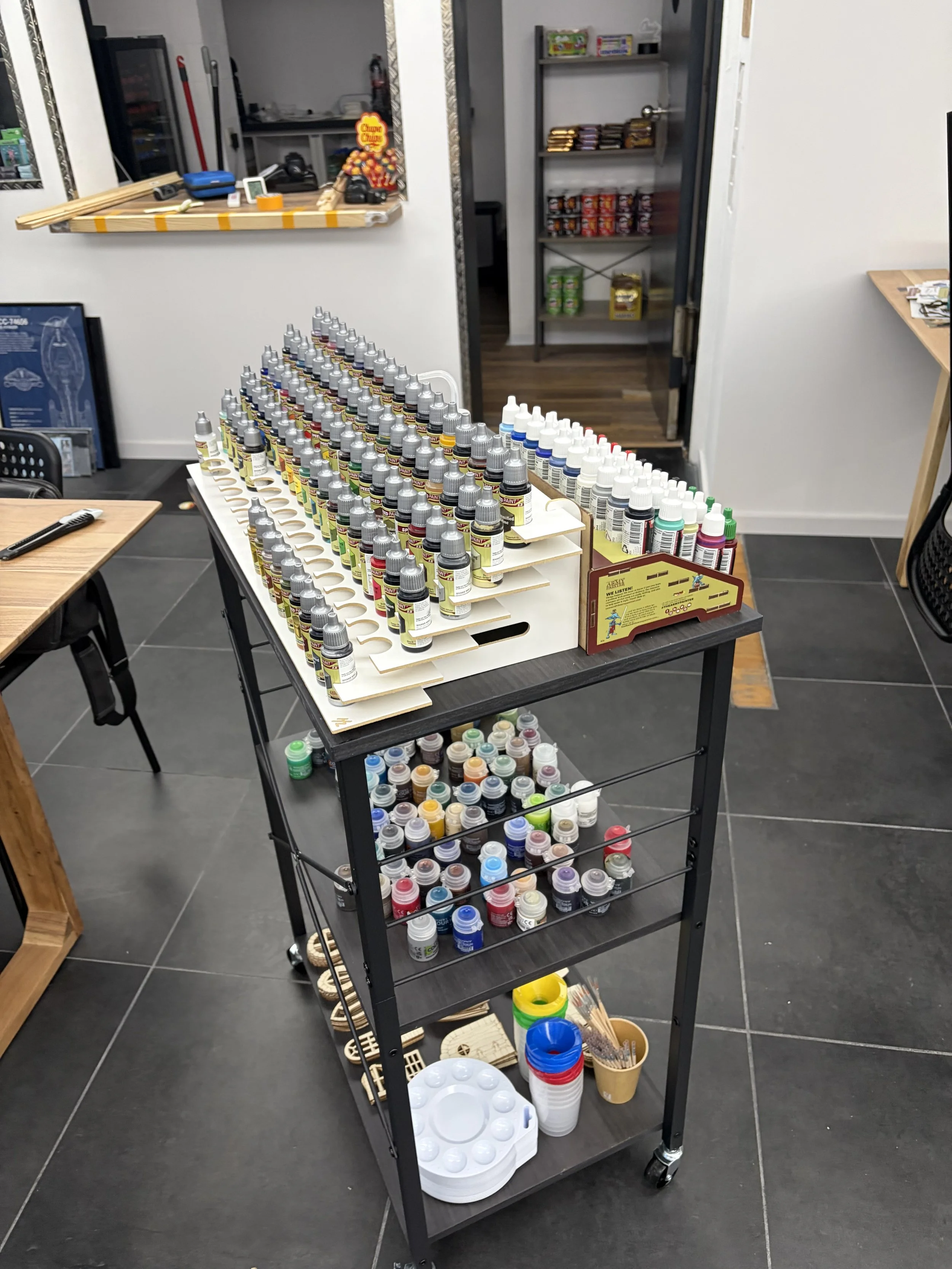 Ein rollendes Regal mit verschiedenen Farbtöpfen und Malmaterialien. Im Hintergrund ein Raum mit Regalen und einem Fenster.