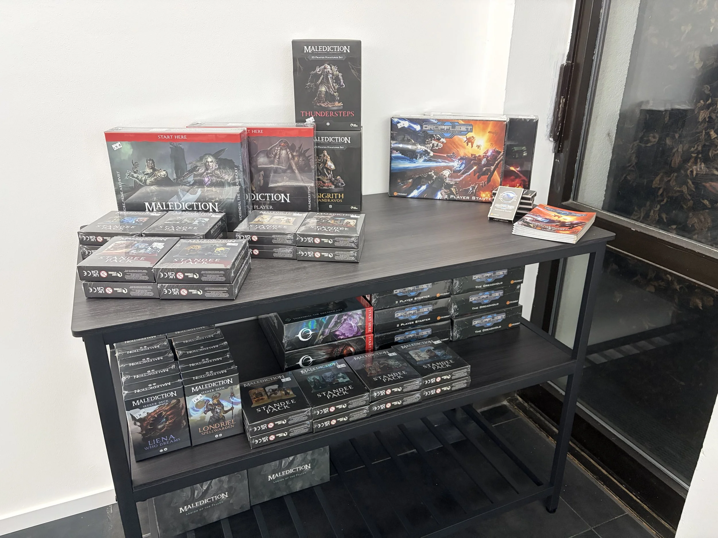 Verschiedene Brettspiele und Miniatur-Sets auf einem schwarzen Regal, darunter 'Malediction', 'Standee Pack' und 'Dropfleet Commander', neben einem Fenster.