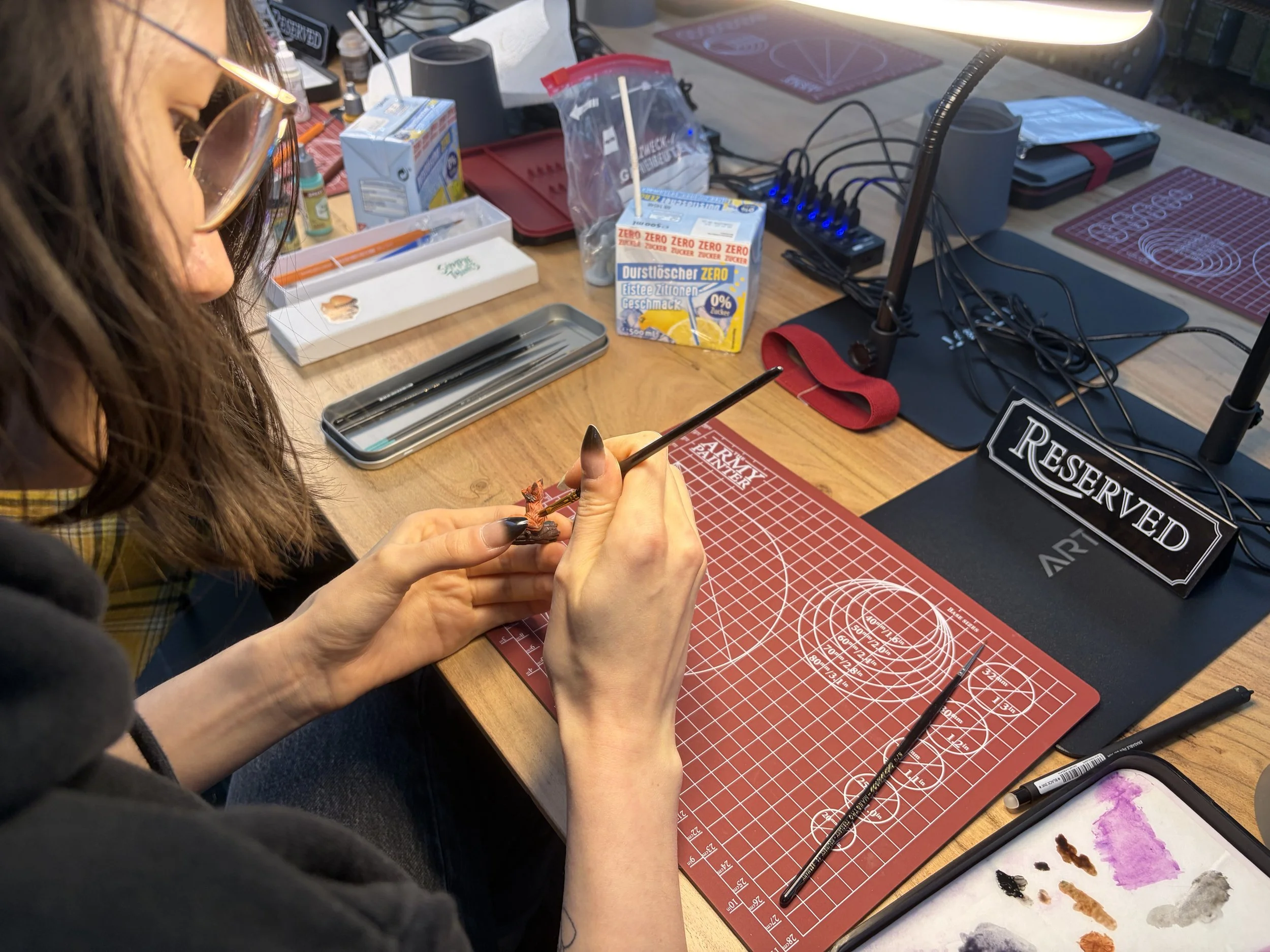 Eine Person mit langen braunen Haaren und Brille arbeitet an einem Modellbauprojekt, während sie an einem Basteltisch sitzt, auf dem Werkzeuge, Getränke und andere Bastelmaterialien liegen.