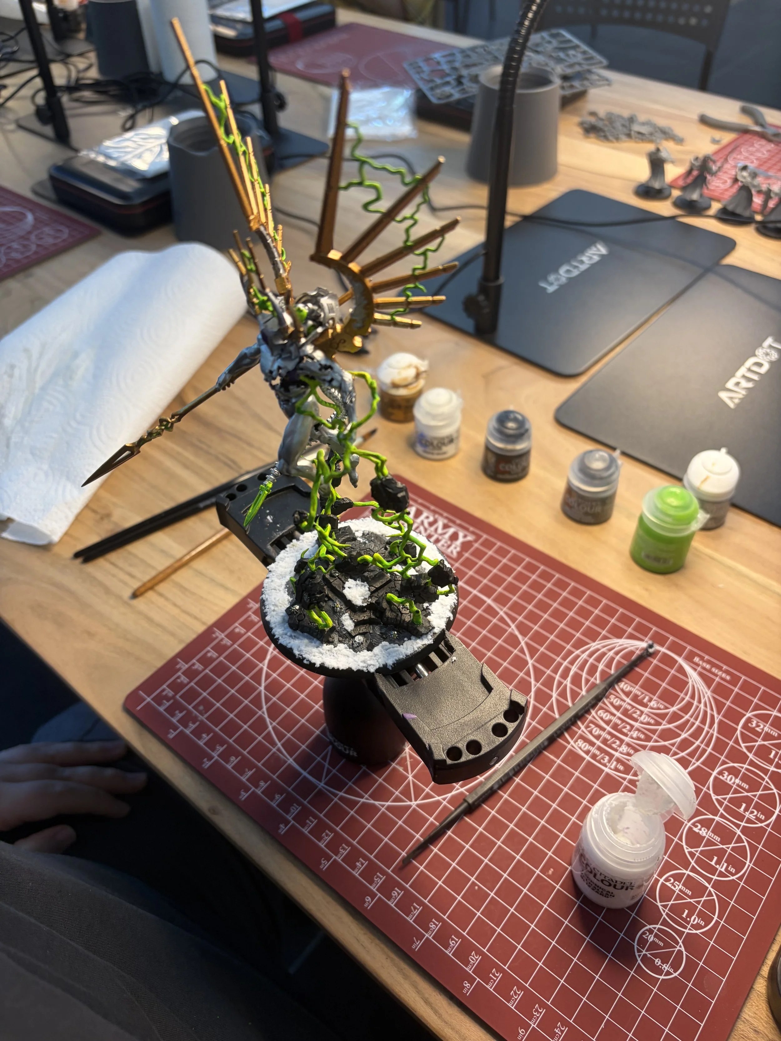 Ein modelliertes Miniaturwesen mit angriffsliedenden, grünen Venen und zerbrochenen, metallischen Armen auf einem schwarzen Stand. Es befindet sich auf einem rot karierten Tisch mit verschiedenen Farben von Farben und Pinsel in der Nähe, sowie Werkze