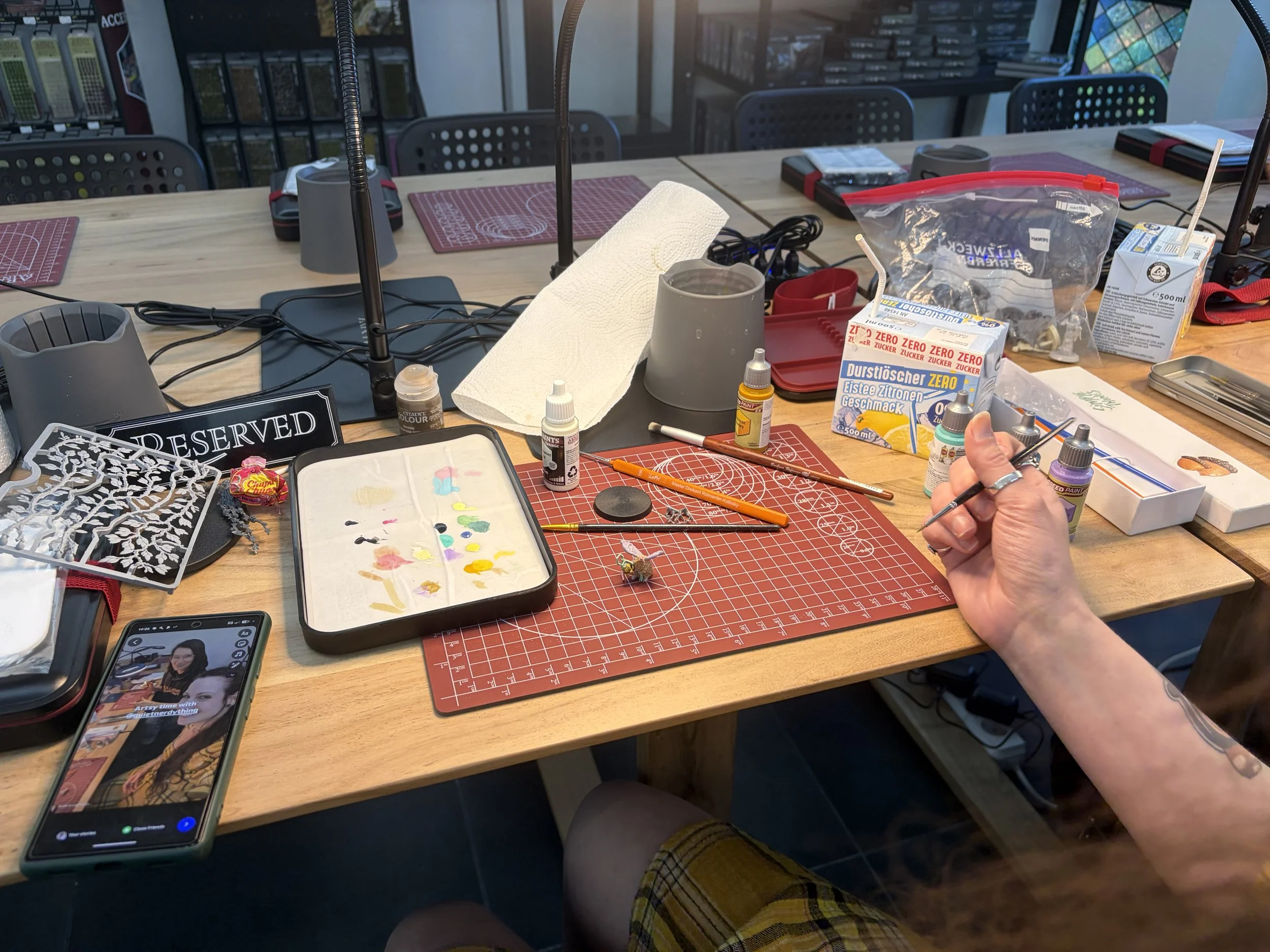 Ein Tisch mit Bastelmaterialien, Farben, Werkzeugen und Dekorationen für eine Bastelaktivität, mit einer Person, die an einem kleinen Details arbeitet.