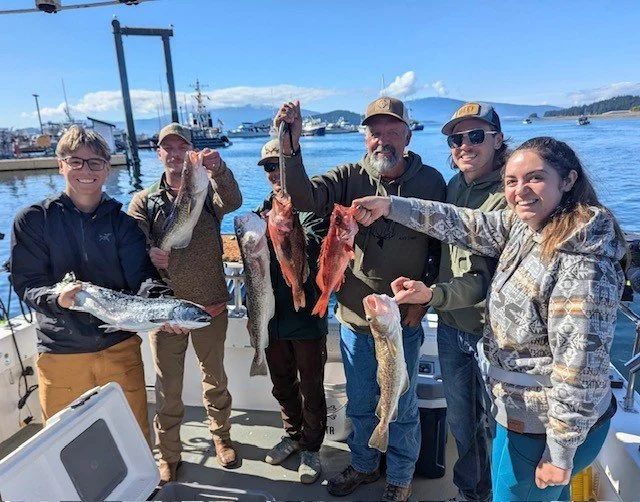 Six people on a boat holding up freshly caught fish with a marina and blue sky in the background.