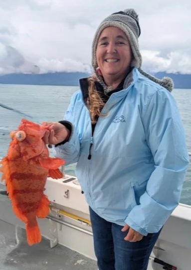 Woman on boat holding a bright orange fish she caught, wearing a blue jacket and gray knit cap, with cloudy sky and water in the background.