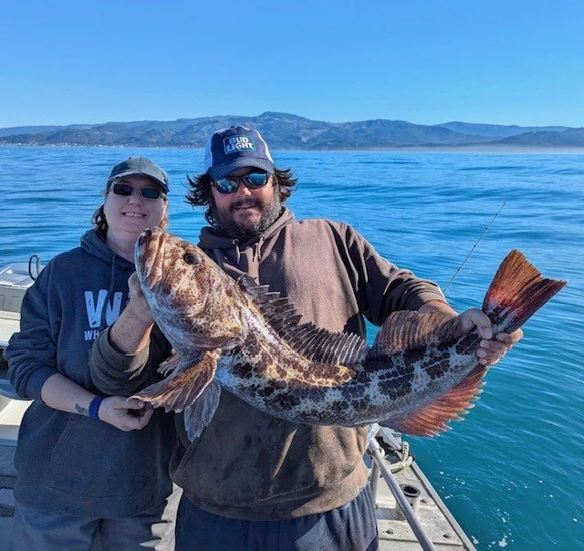 Two people holding a large fish on a boat with Pacific ocean and distant mountains in the background.