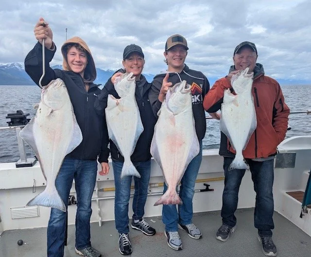 Four people on a boat holding large flatfish they caught, with the ocean and cloudy sky in the background.